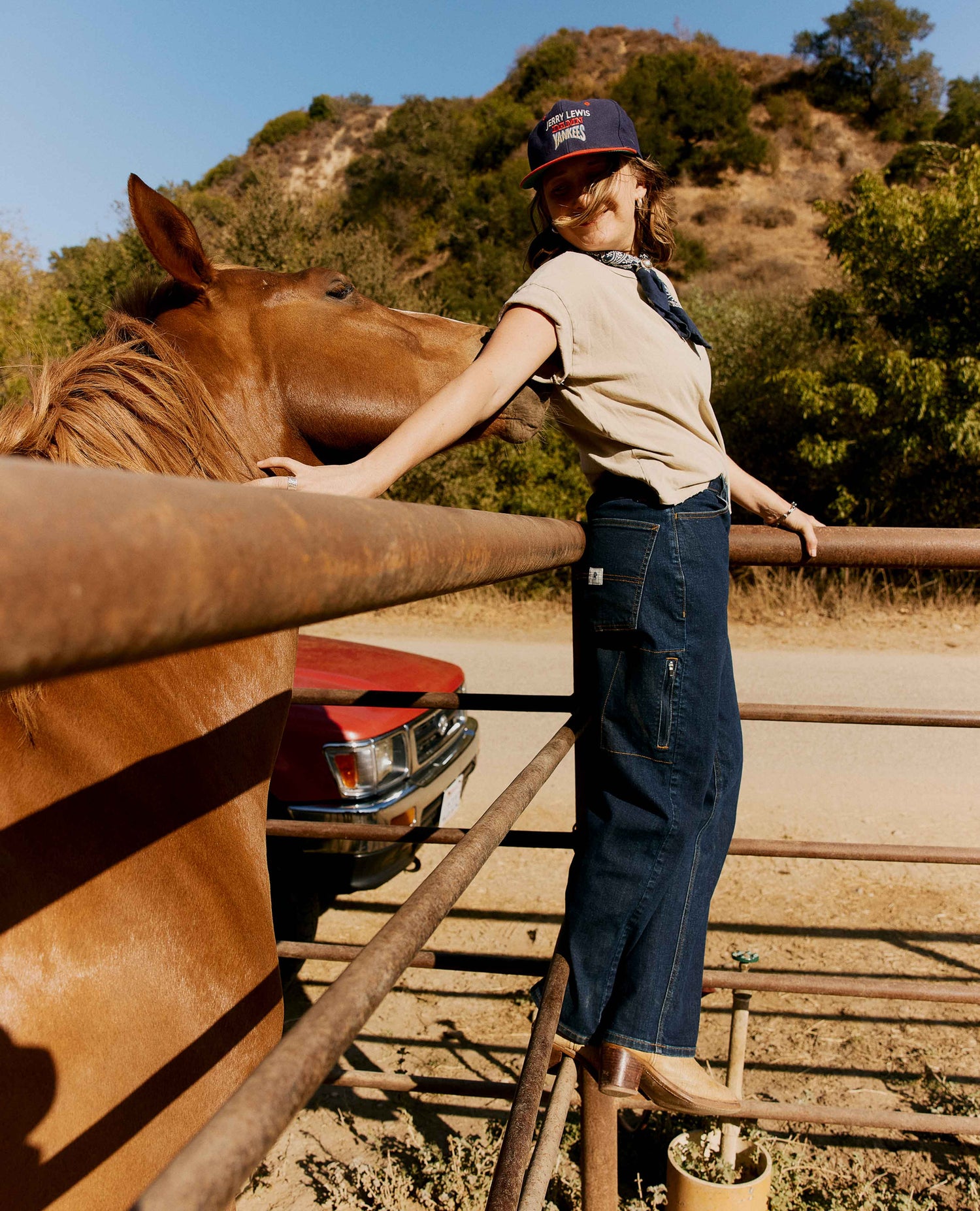 A woman stands on metal rails beside a horse in an outdoor ranch setting, wearing a beige sleeveless top, dark blue wide-leg jeans, and a navy baseball cap. A horse is visible on the left, with a red vehicle and dry hillside in the background.