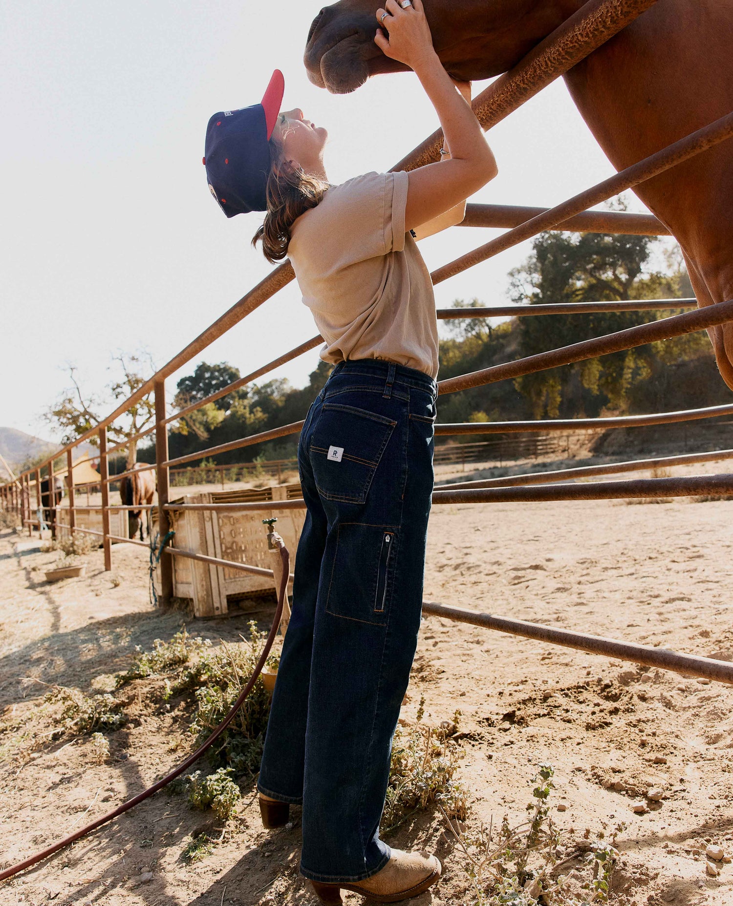 A woman in a beige t-shirt, dark blue wide-leg denim jeans, and a red-and-black cap stands in a sunlit horse paddock, reaching up to touch a horse's face. The background shows a sandy paddock with metal rails and trees under a bright sky.