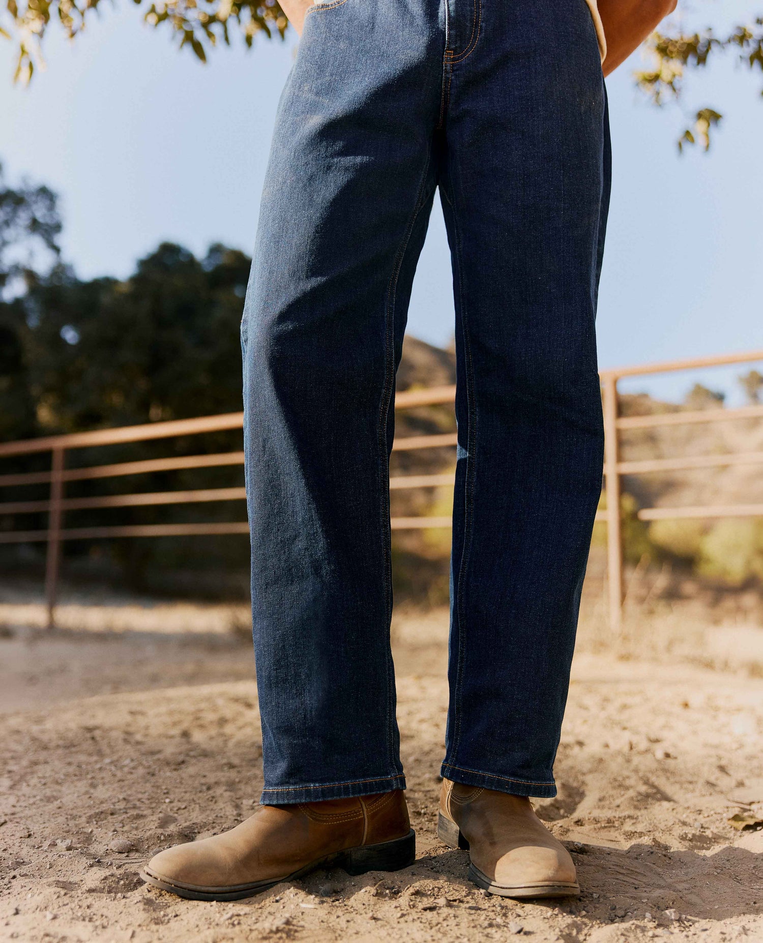 Close up of a man's legs in blue jeans and cowboy boots, standing outside in front of a fence. 