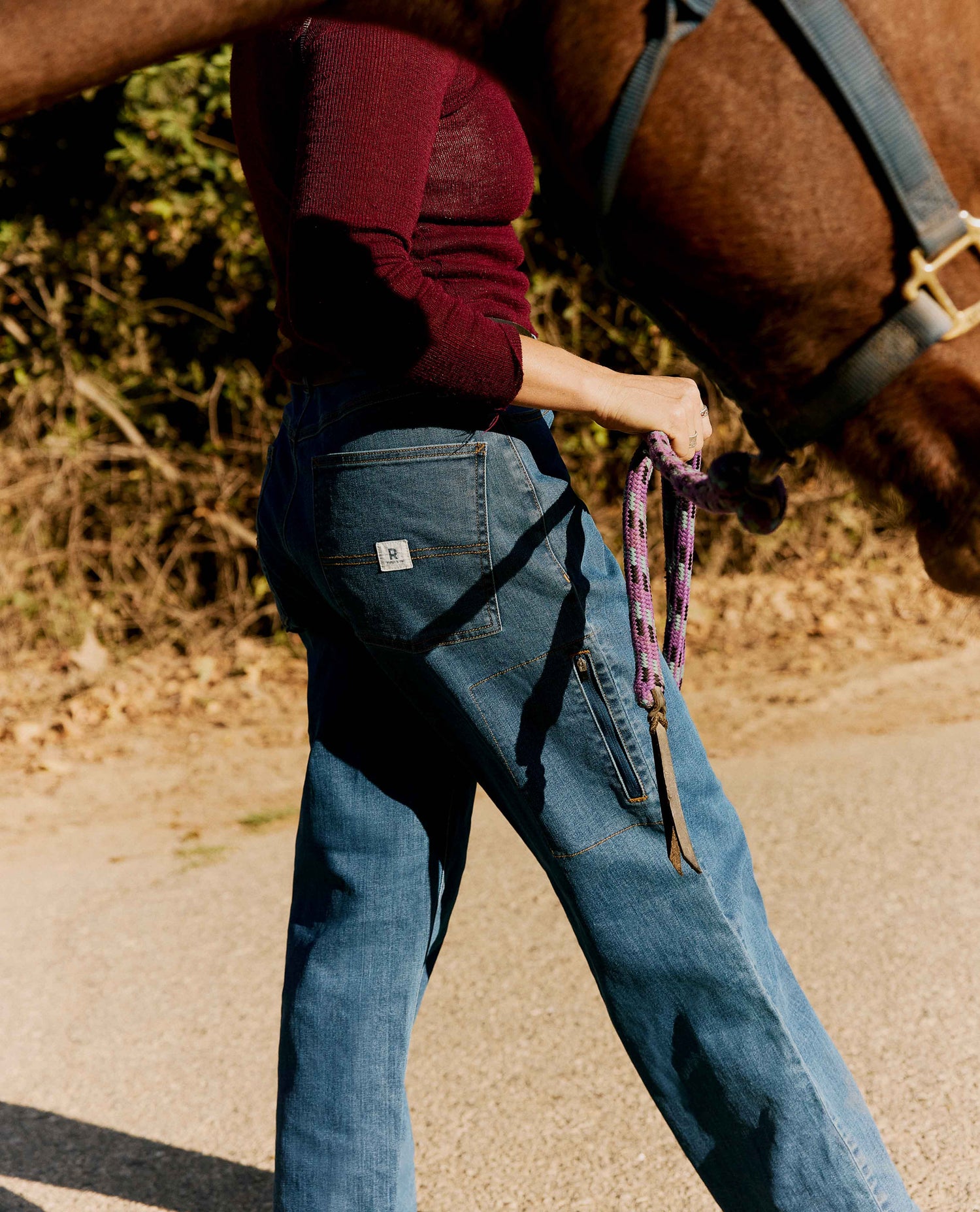 A person wearing blue denim jeans and a burgundy long-sleeve top walks alongside a brown horse outdoors on a sunlit road. The jeans have a small white brand patch on the back pocket.