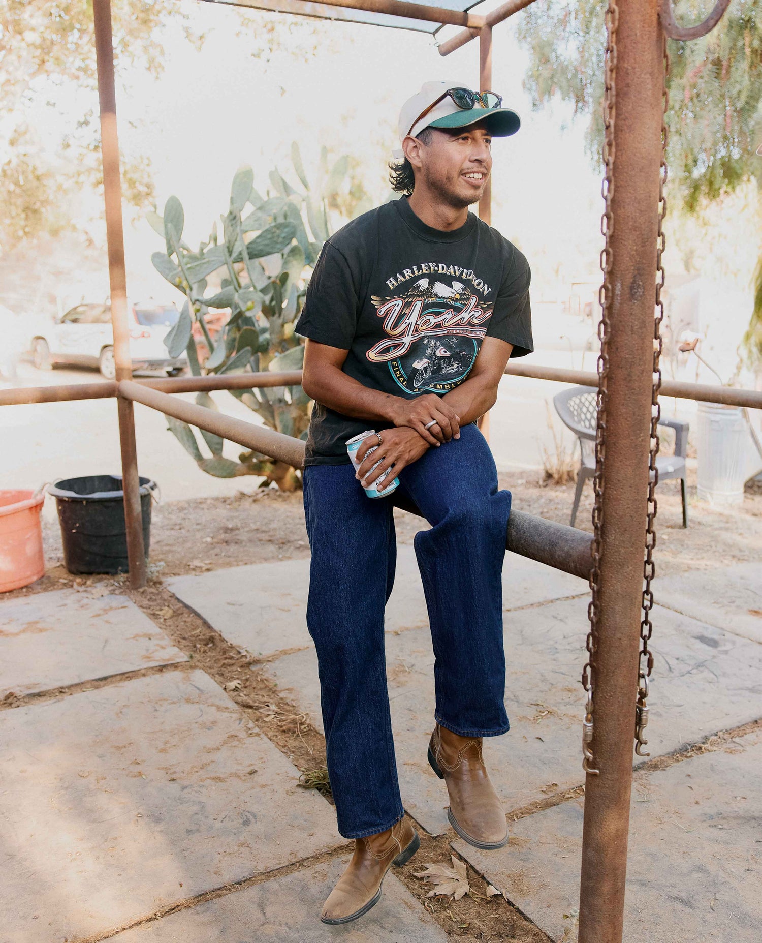 Man sitting on fence, wearing blue jeans, cowboy boots, and a black shirt.