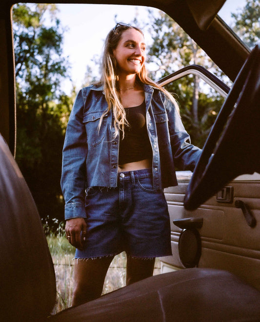 A woman smiling and looking out from inside a vehicle, wearing a cropped blue denim shirt and denim shorts over a black crop top.