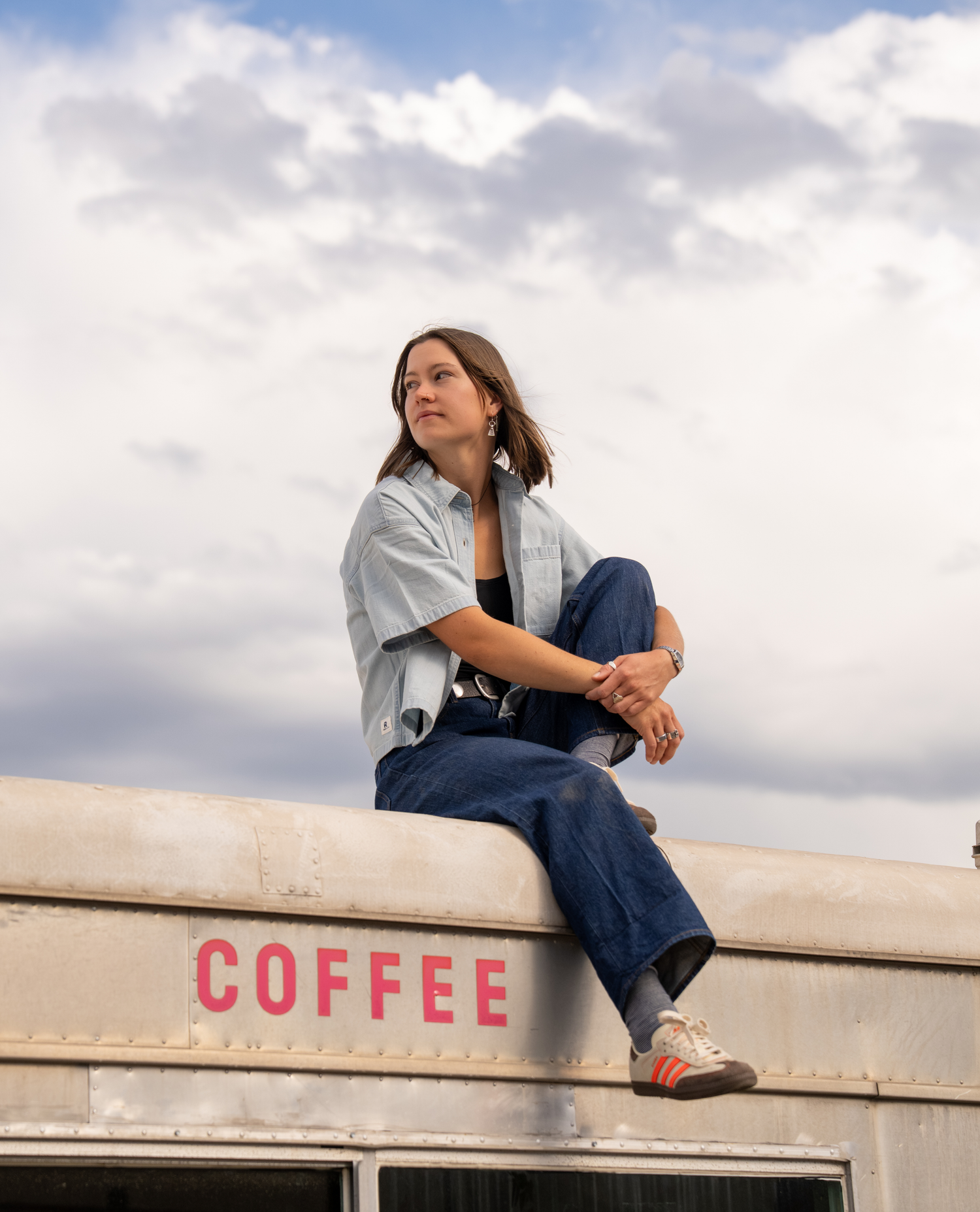 A woman sits on the roof of a vintage-style bus under a cloudy sky, wearing a light blue button-up shirt, dark blue jeans, and white sneakers with orange stripes. The side of the bus has the word 'COFFEE' in red letters.