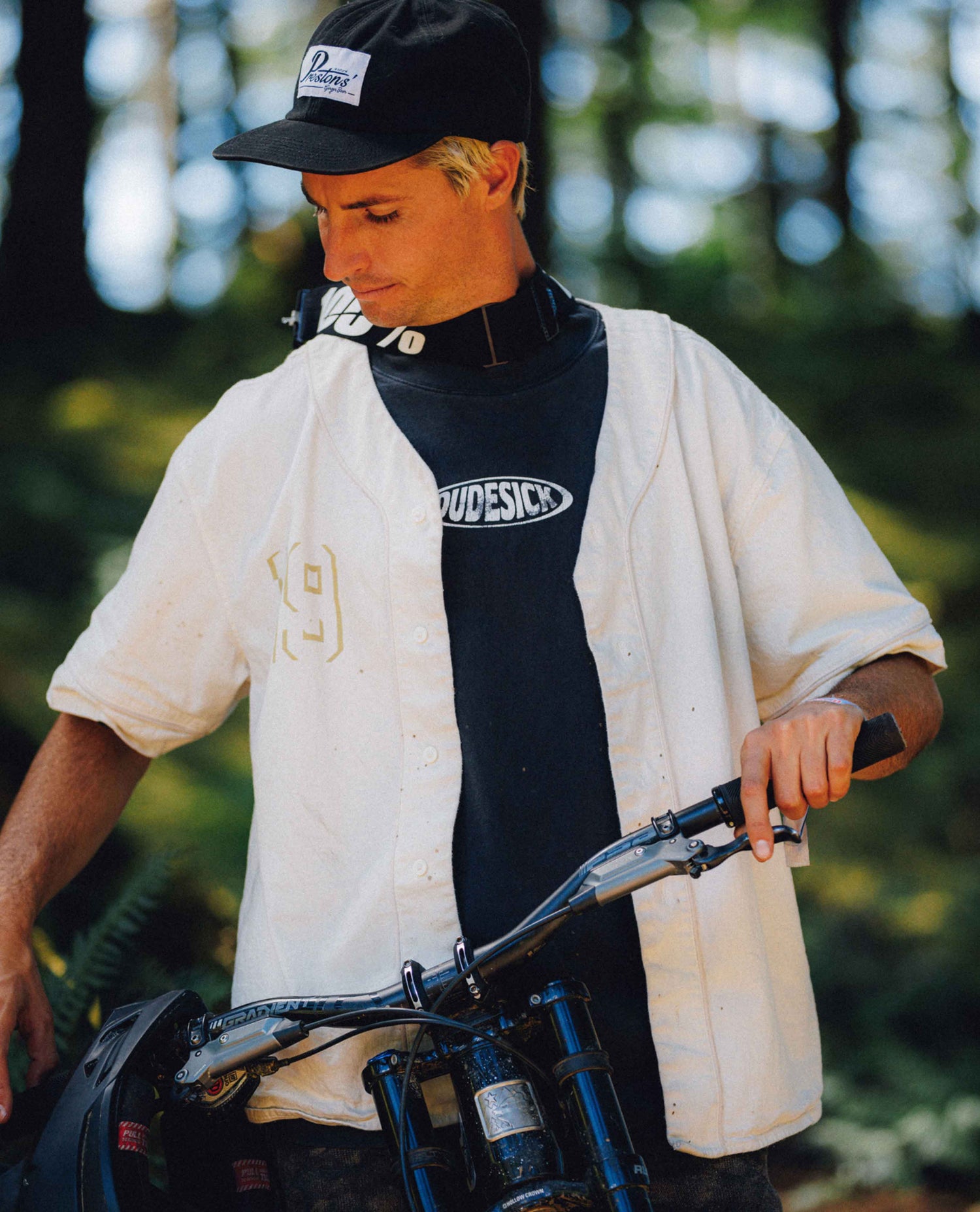 A man wearing a black cap, a white short-sleeve button-up shirt with a gold '19' on the chest, and a black t-shirt with 'WIDESCS' printed on the front, holding the handlebars of a blue mountain bike in an outdoor forest setting.