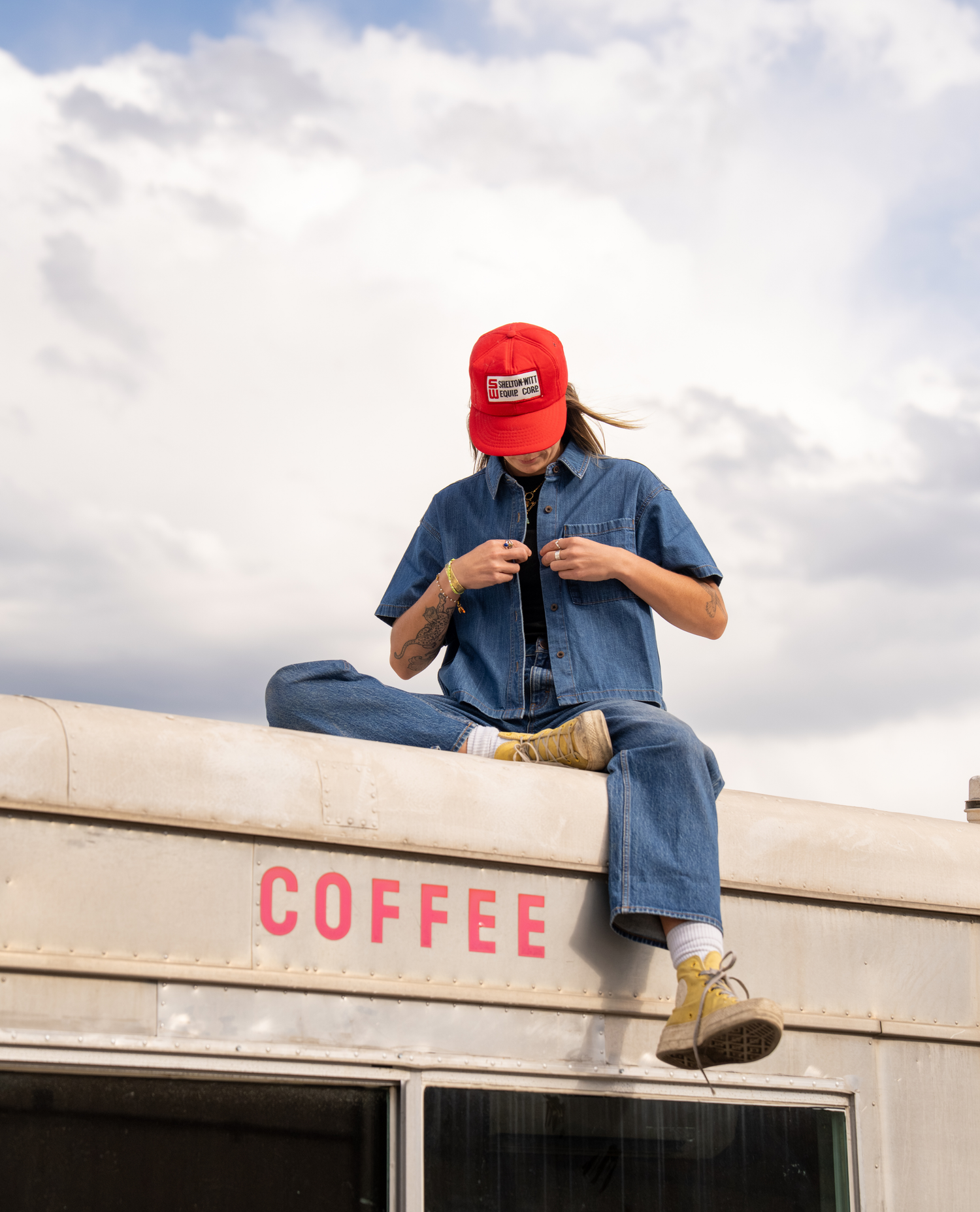 A person sits on the roof of a vintage bus under a cloudy sky, wearing a red baseball cap, a blue denim shirt and jeans, and tan boots. The bus has the word 'COFFEE' printed in red on the side.