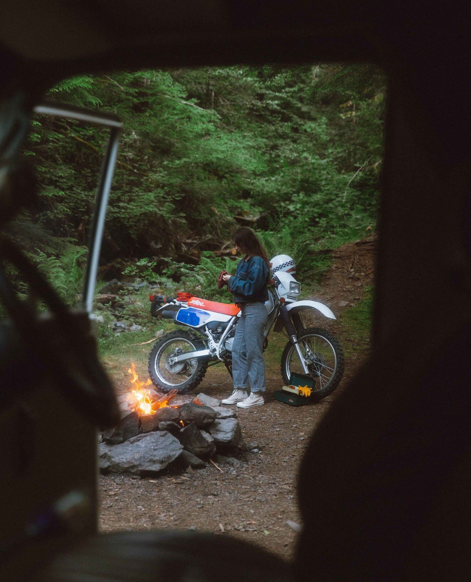 Photo taken from inside a car looking out at alex by the campfire with her dirt bike