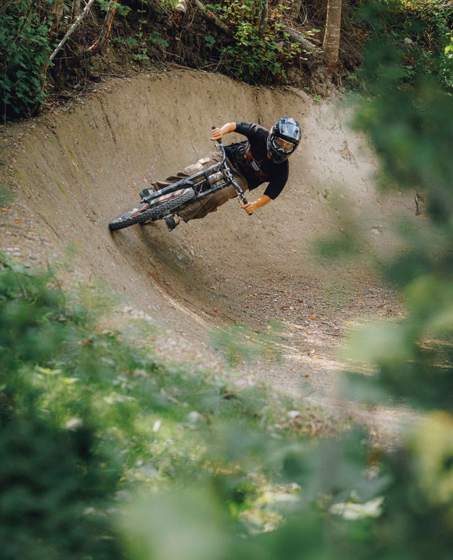 A mountain biker in a black shirt and helmet leans sharply into a dirt ramp on a forest trail, with blurred green foliage in the foreground.