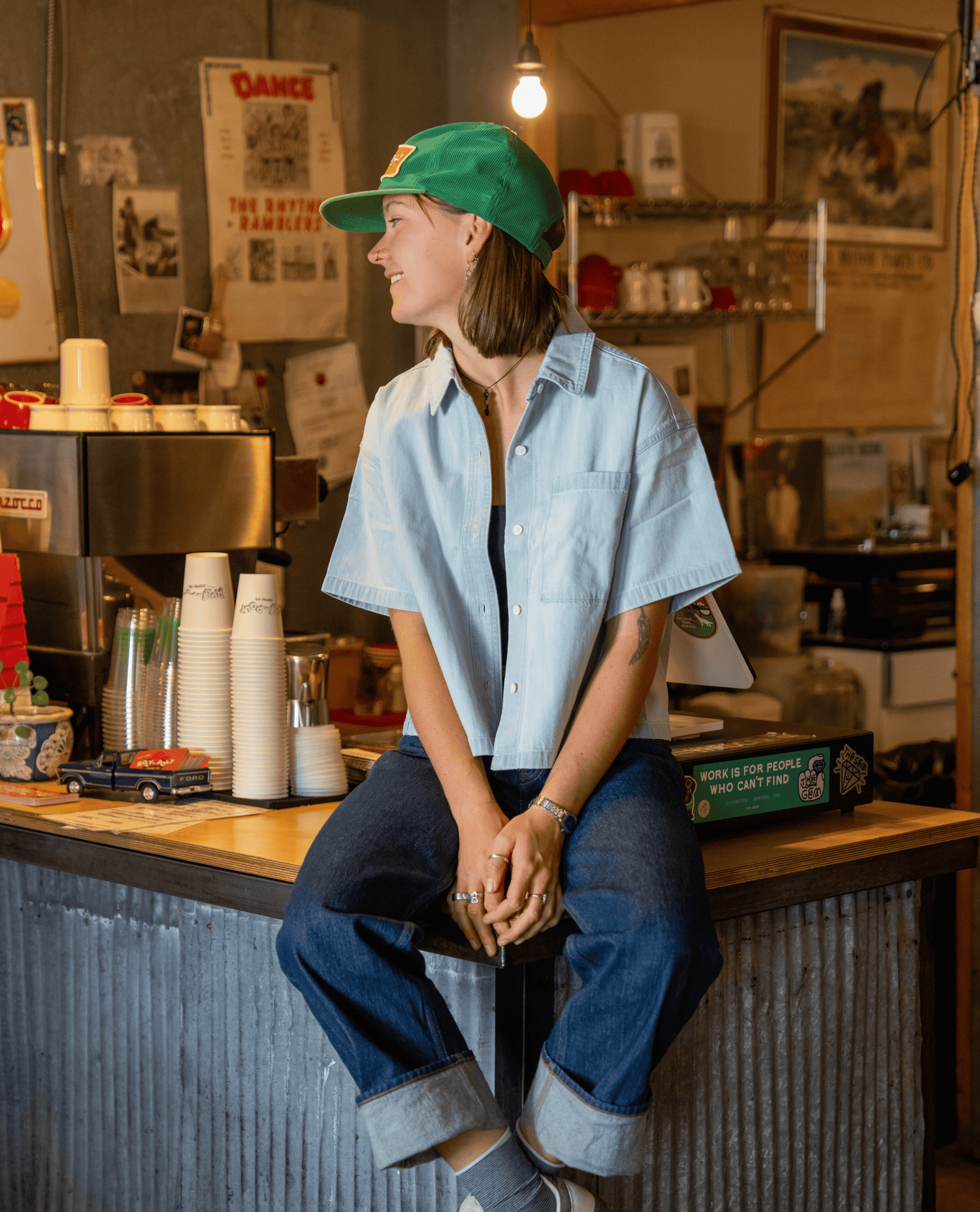 A woman sits on a stool behind a coffee counter wearing a light blue cropped button-up shirt, dark blue jeans with rolled cuffs, a green baseball cap, and gray socks in a cozy, vintage-style coffee shop.