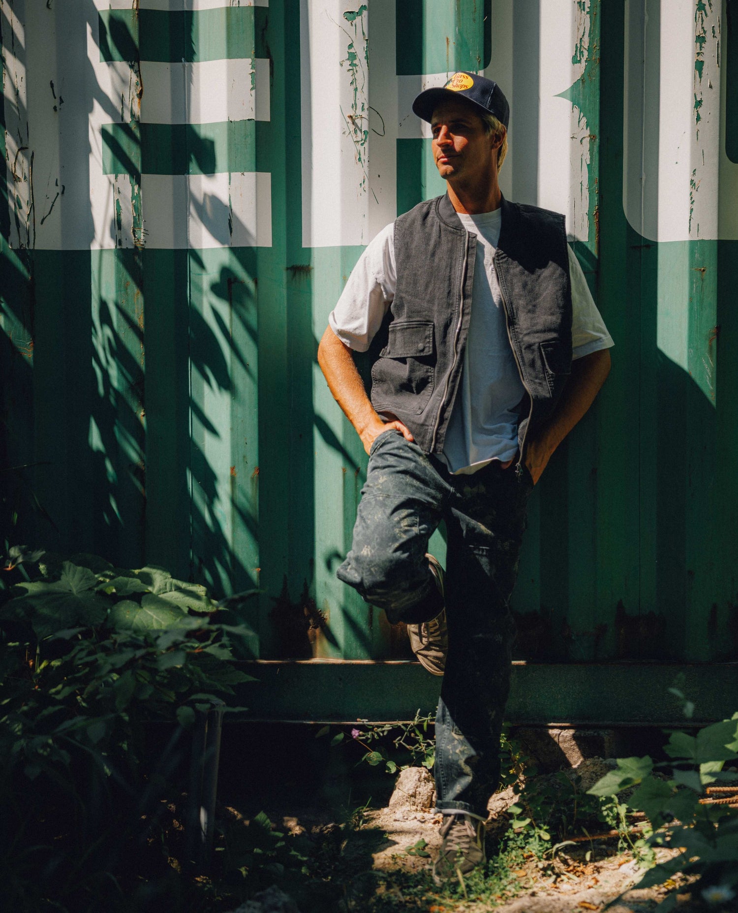 A man wearing a dark gray utility vest over a white t-shirt, dark jeans, and a baseball cap leans against a weathered green and white corrugated metal wall outdoors.