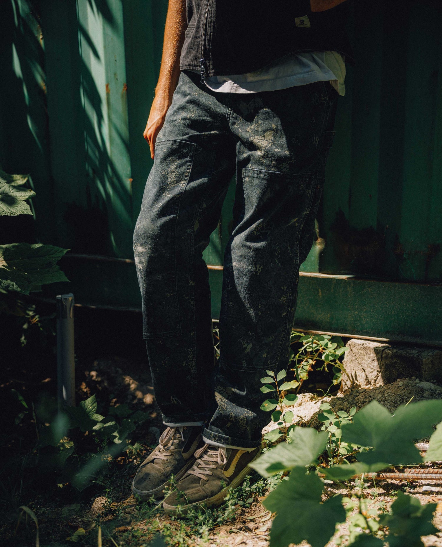A person wearing dark, heavily distressed denim jeans with a white t-shirt and brown boots, standing outdoors in front of a green wooden fence with leafy plants in the foreground.