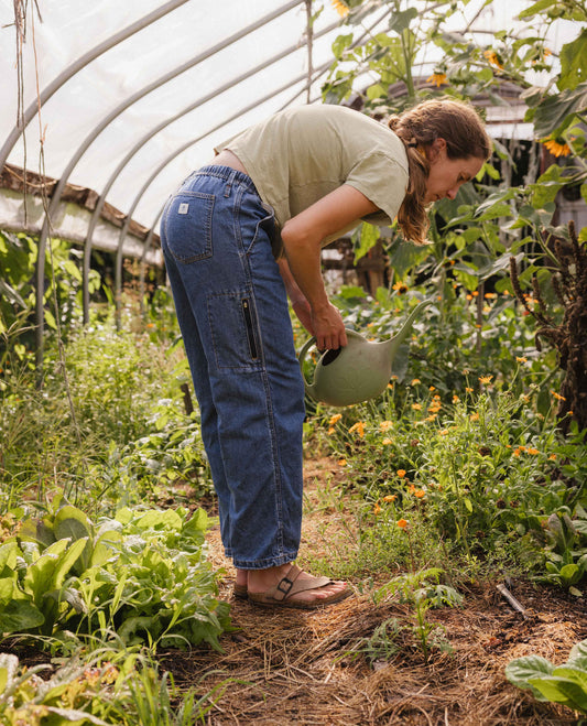A woman watering plants in a greenhouse wearing blue denim cargo pants, a light olive green t-shirt, and brown sandals.
