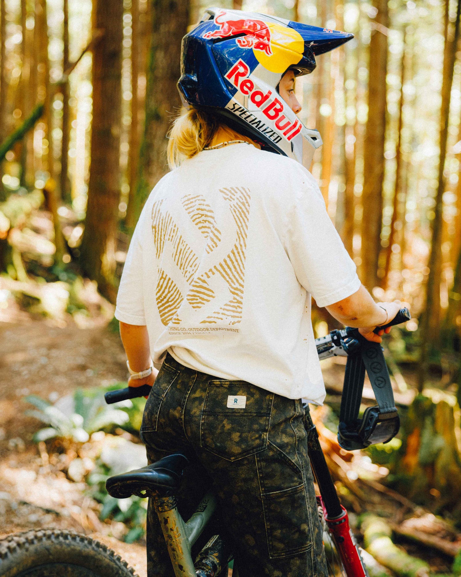 A person wearing a white graphic t-shirt with a gold back design, camouflage pants, and a Red Bull helmet rides a mountain bike on a forest path.