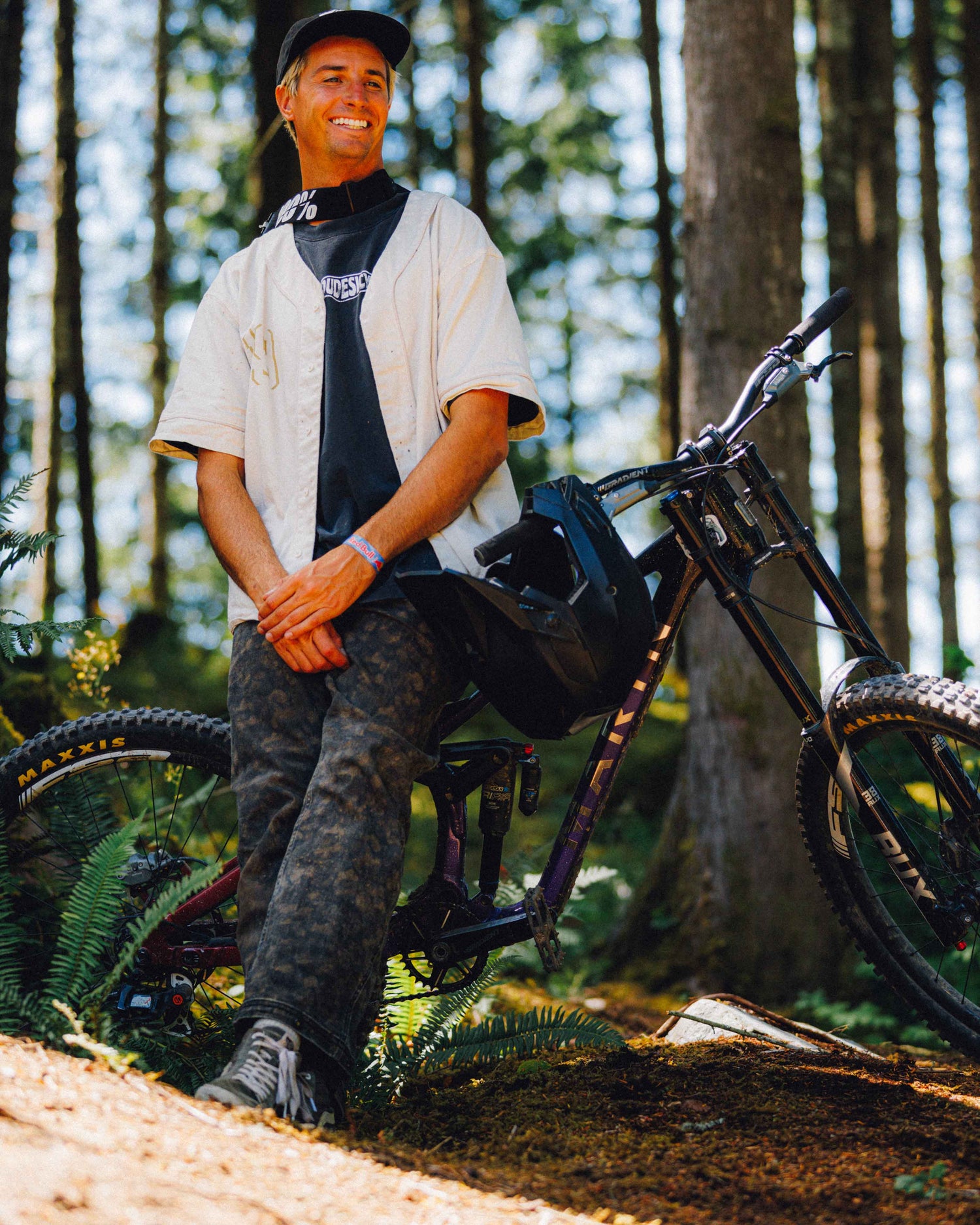 A man wearing a black cap, a white jersey over a black shirt, and camouflage pants sits on a mountain bike in a forest setting.