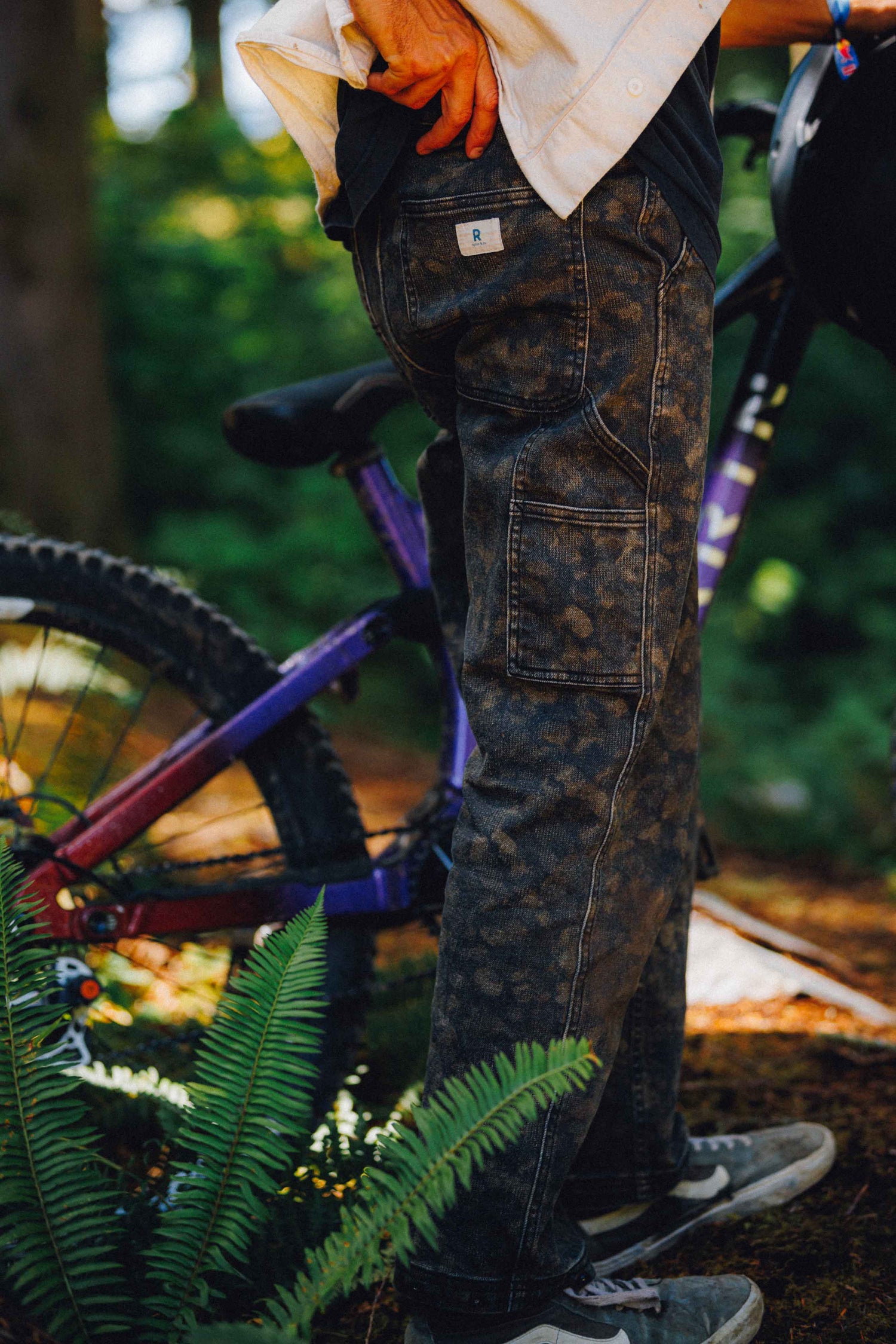A person outdoors wearing dark camouflage cargo pants, standing next to a purple mountain bike with green ferns in the foreground.