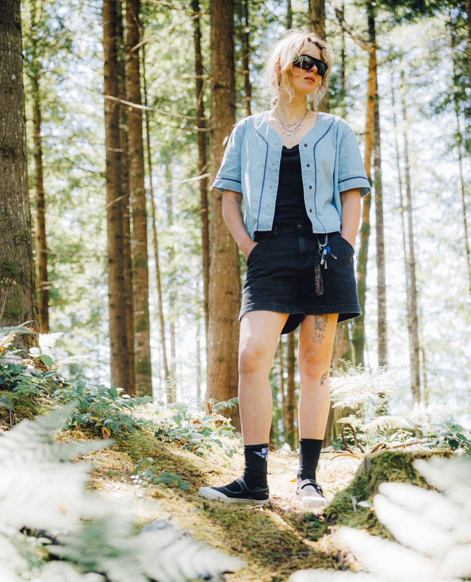 A woman stands in a sunlit forest wearing a light blue cropped button-front shirt, a black mini skirt, a black top underneath, black socks, and black sneakers.
