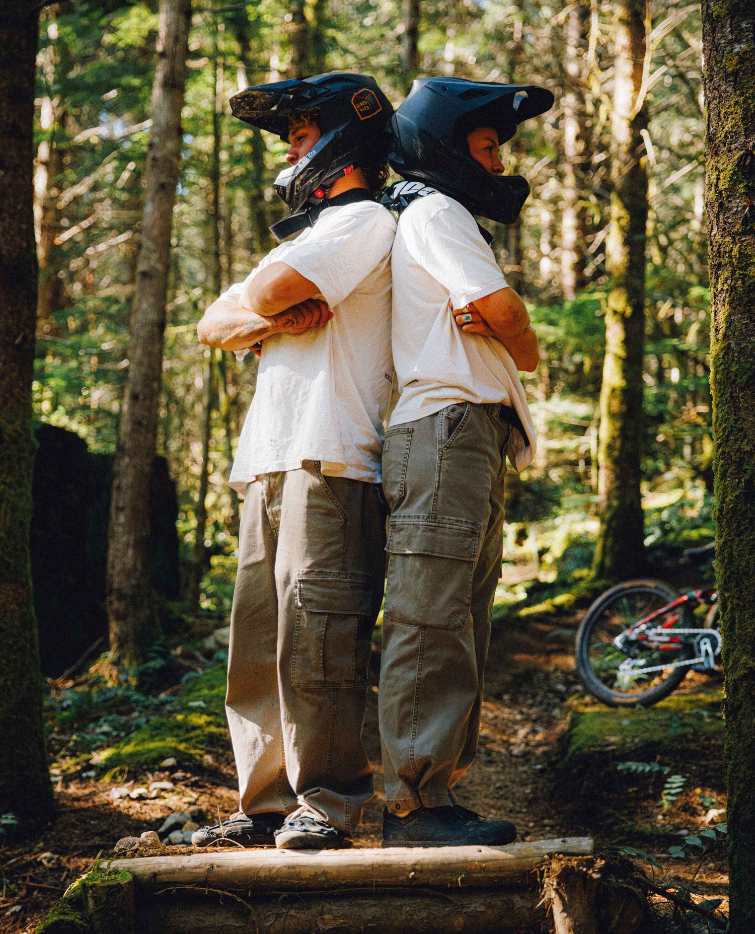 Two people wearing white t-shirts, cargo pants, and black helmets stand back-to-back on a forest trail, with a mountain bike visible in the background.