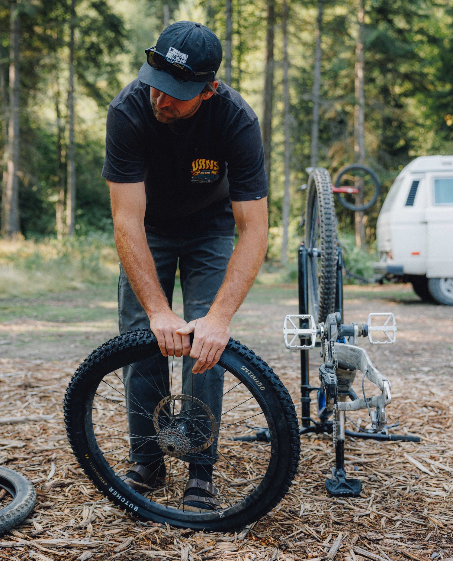 A man in a black t-shirt and cap stands outdoors on a wooded trail next to a bicycle frame and wheel, holding a black knobby tire.