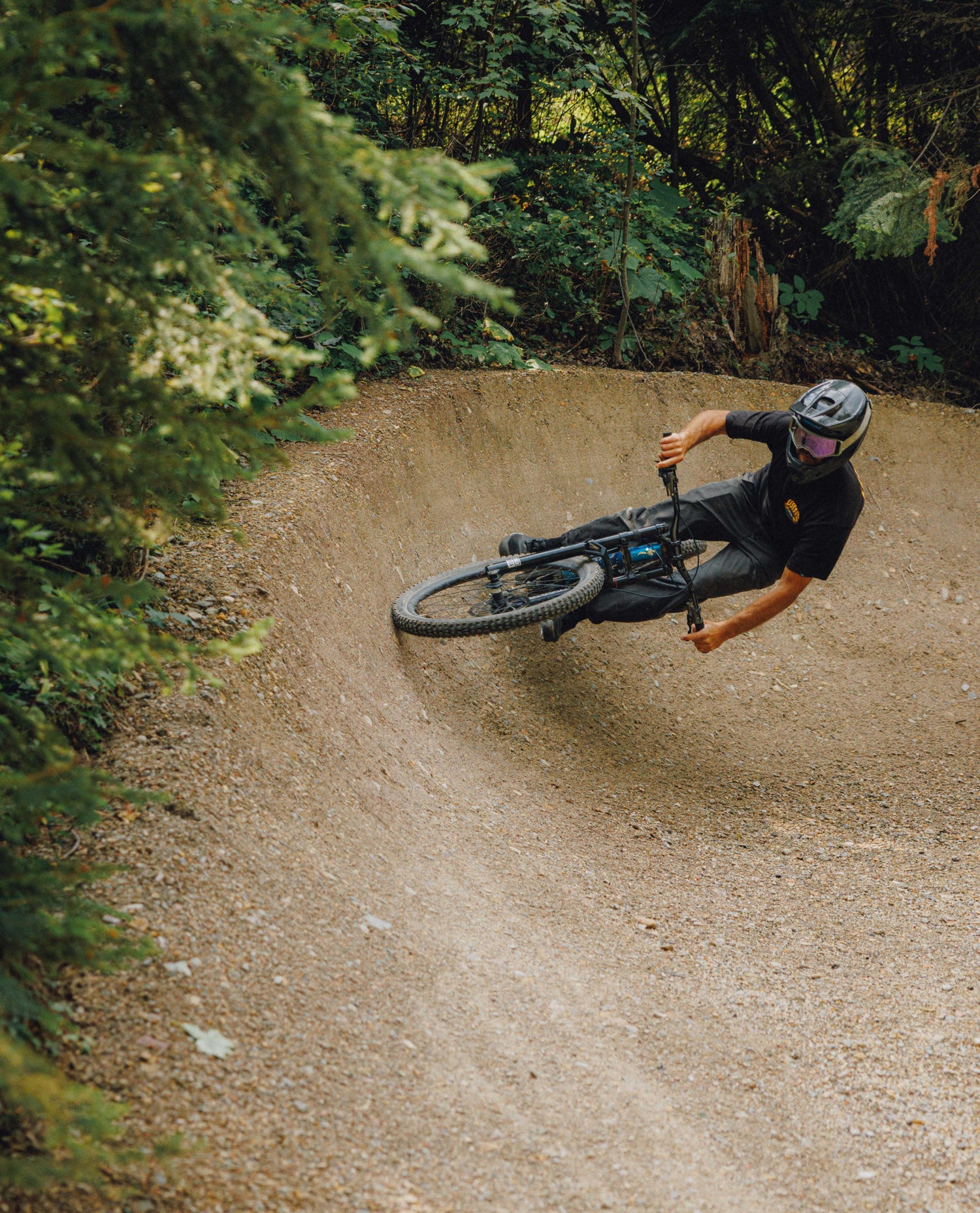 A mountain biker in a black shirt, gray pants, and a black helmet with purple-tinted goggles leans sharply into a turn on a dirt trail surrounded by dense green forest.
