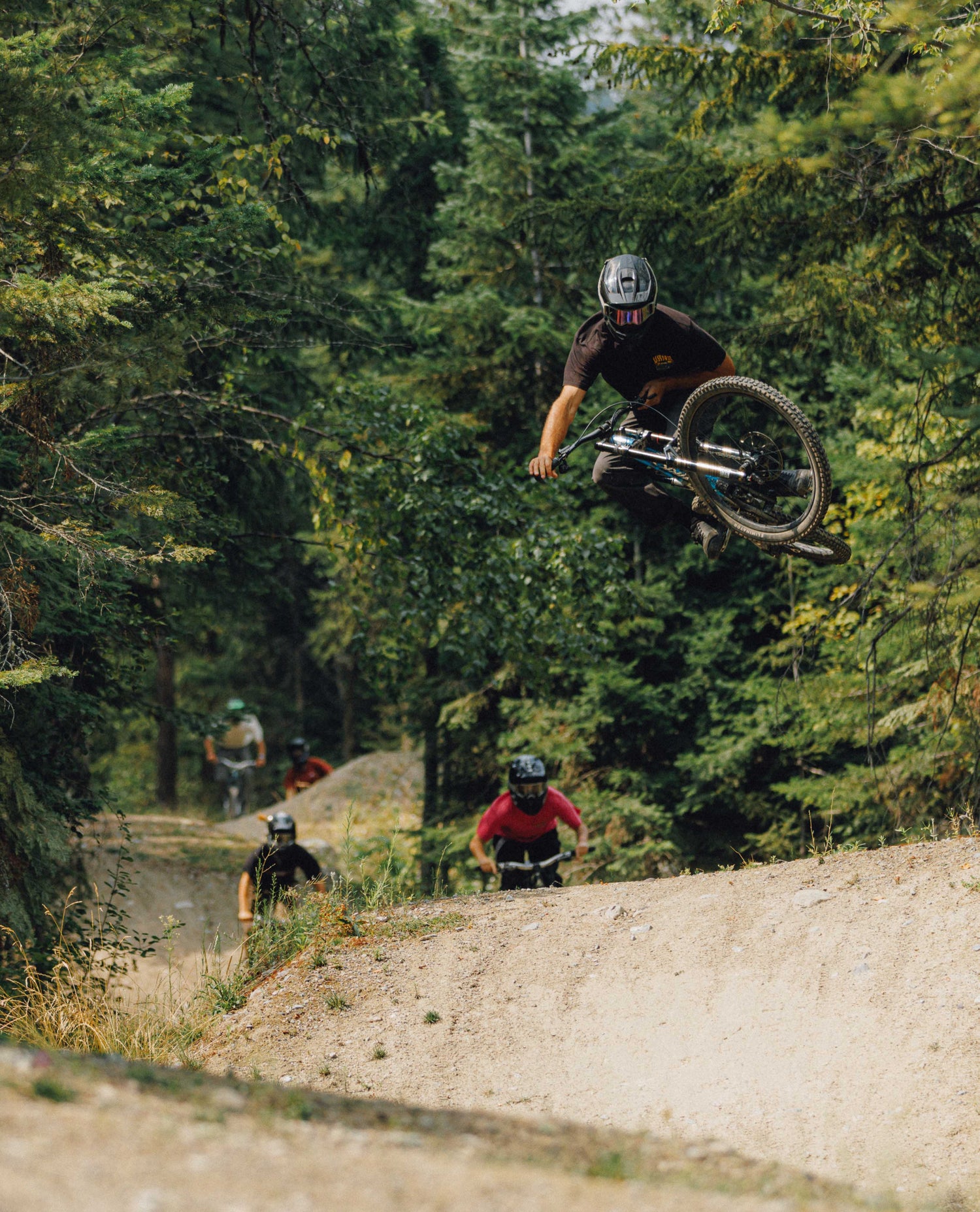 Mountain biker mid-air on a dirt trail surrounded by dense green forest, with other riders visible below on the trail.