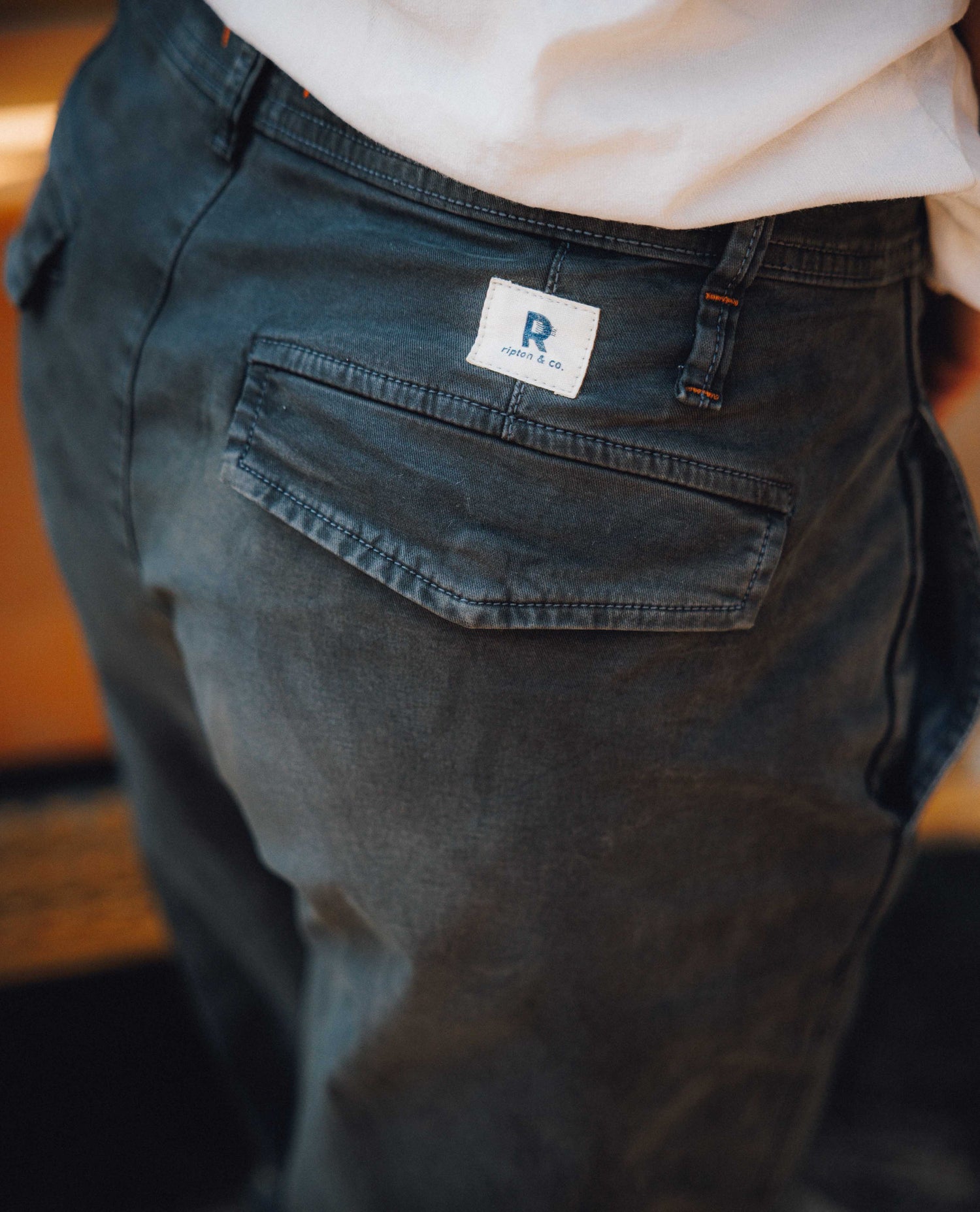 Close-up back view of a person wearing dark gray washed denim jeans with a white brand patch on the back pocket.