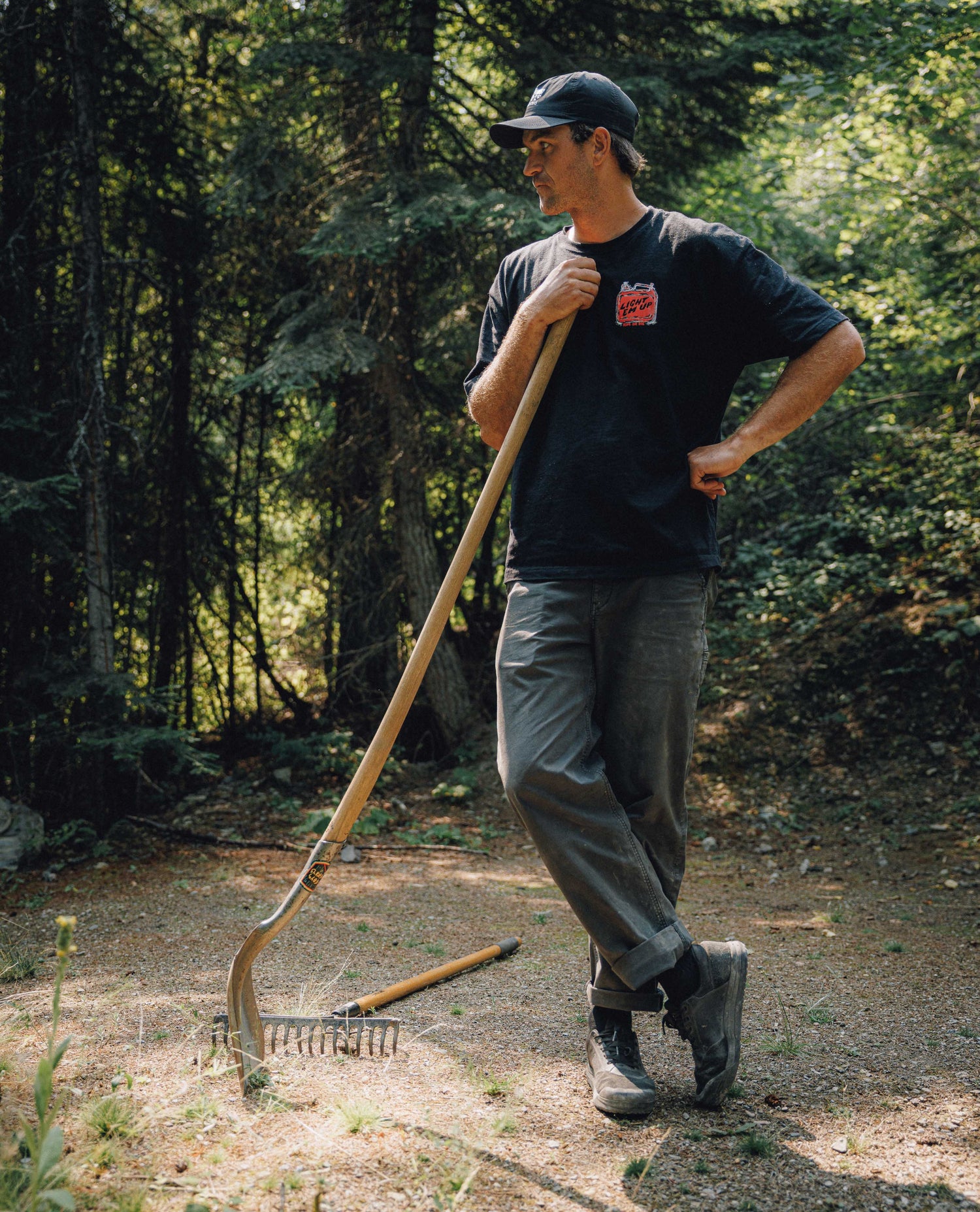 A man stands in a wooded outdoor setting holding a long-handled rake. He is wearing a black t-shirt with a red graphic on the chest, dark gray pants, and a black baseball cap.