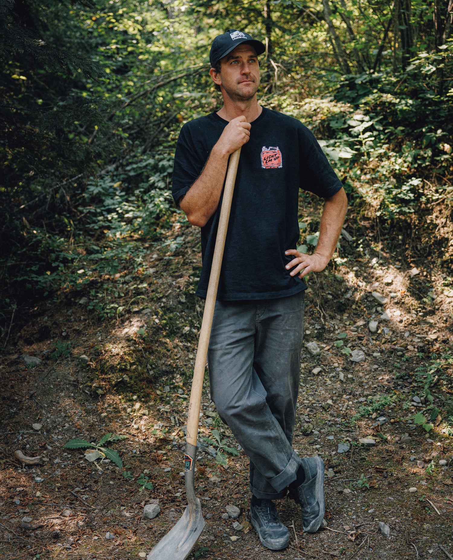 A man standing on a wooded trail holding a shovel, wearing a black t-shirt with a red and white graphic on the left chest, gray pants, and a black cap.