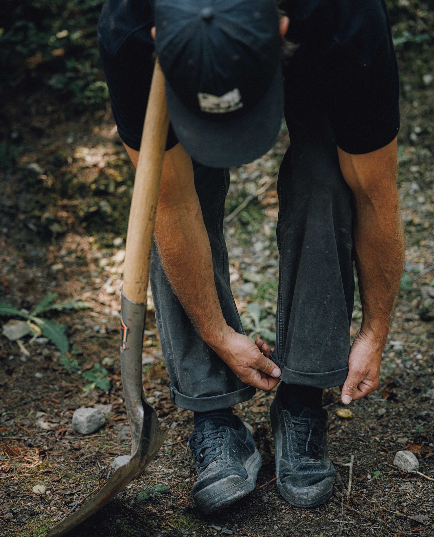 A person outdoors in a wooded setting, bending down to adjust the cuffs of dark gray work pants while holding a shovel. The person is wearing a black t-shirt, a black baseball cap, and black work boots.