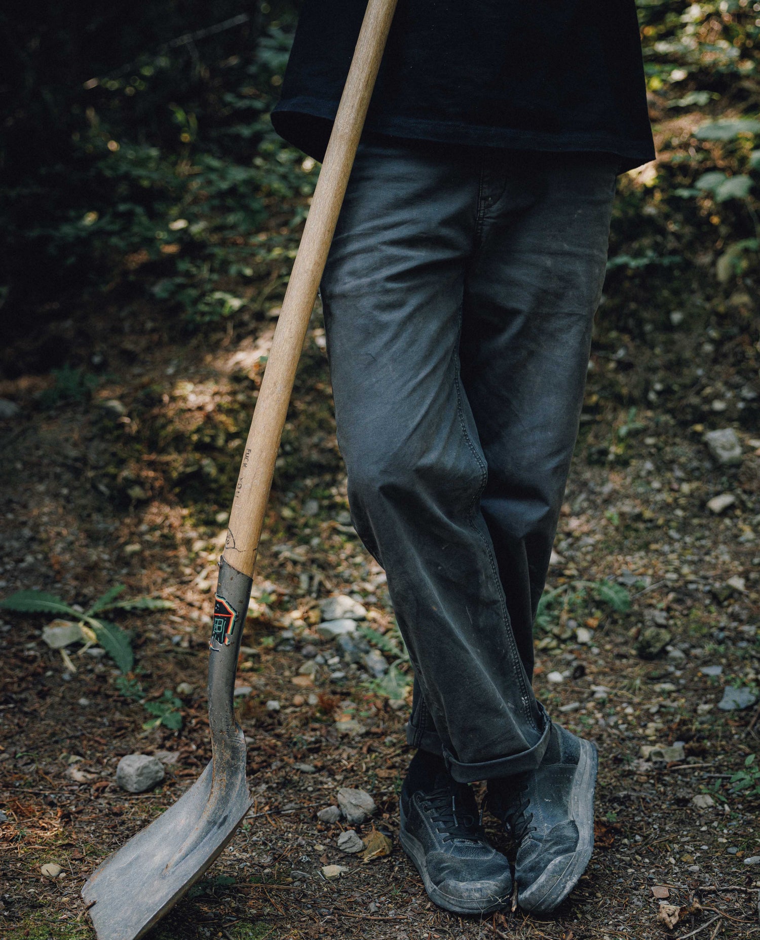A person standing outdoors on a wooded path wearing dark gray work pants and black work boots, holding a shovel with a wooden handle.