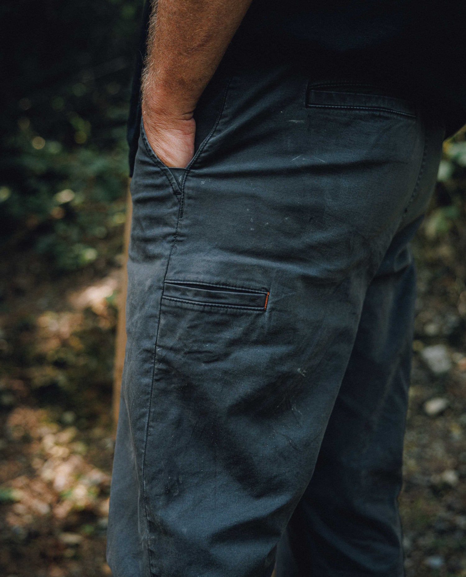 Close-up of a person wearing dark charcoal gray work pants with a side pocket and a small orange tab, standing outdoors in a wooded setting.