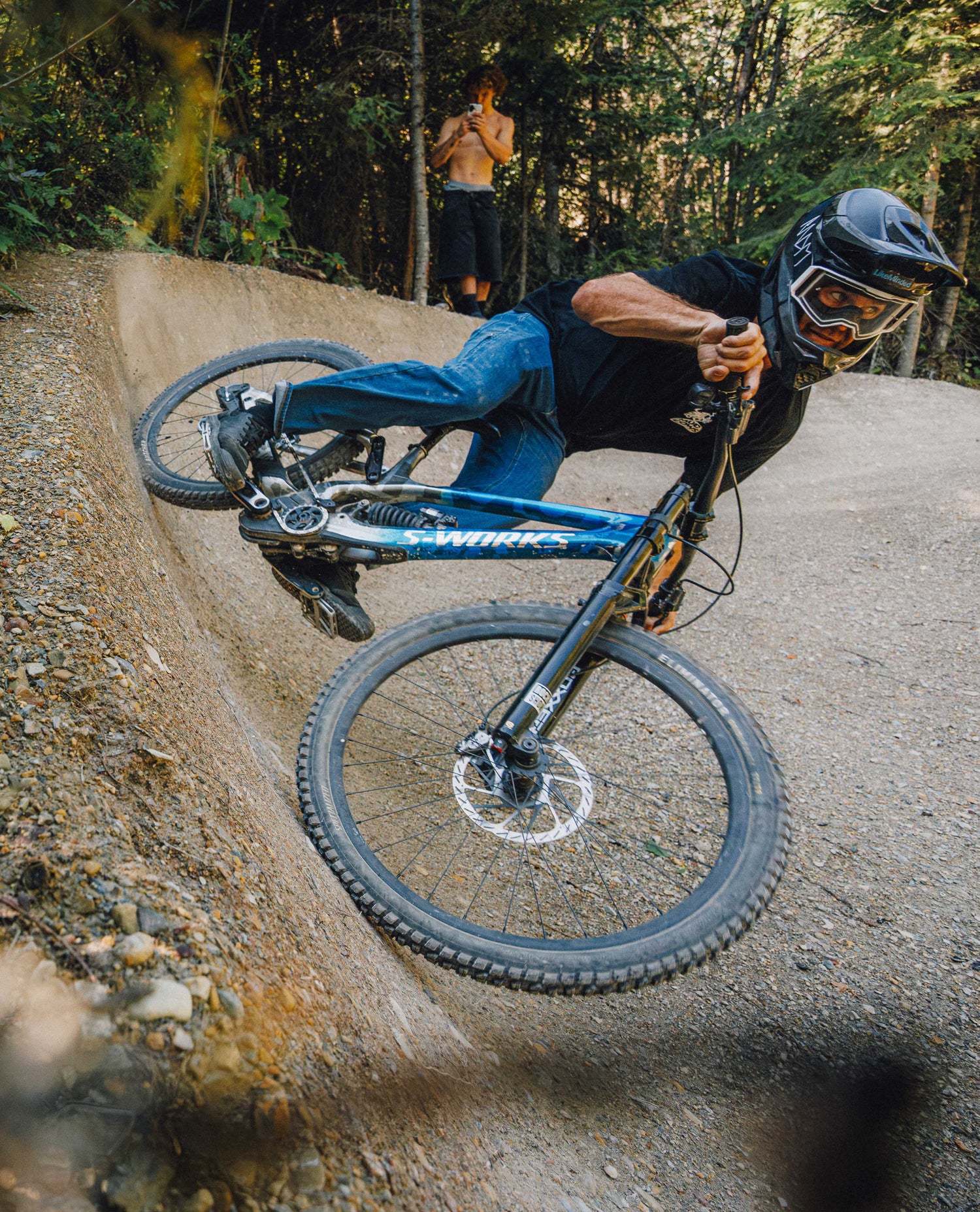A mountain biker in a black helmet and black shirt rides a blue and black Specialized full-suspension bike around a dirt ramp in a forest setting.