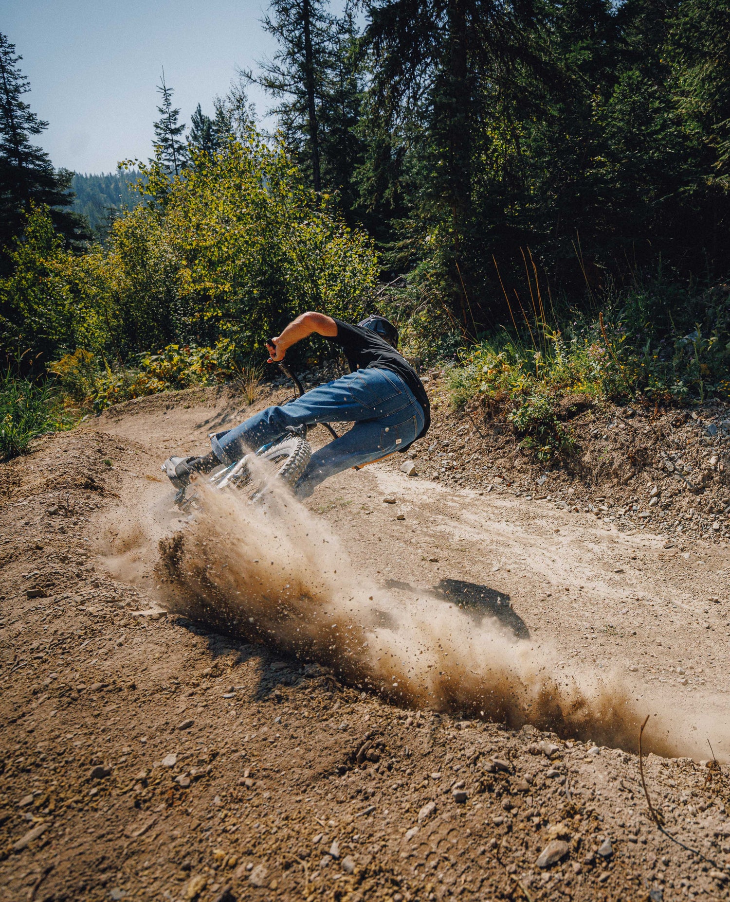 A mountain biker leans sharply into a turn on a dirt trail, kicking up a large cloud of dust, with dense green forest in the background.