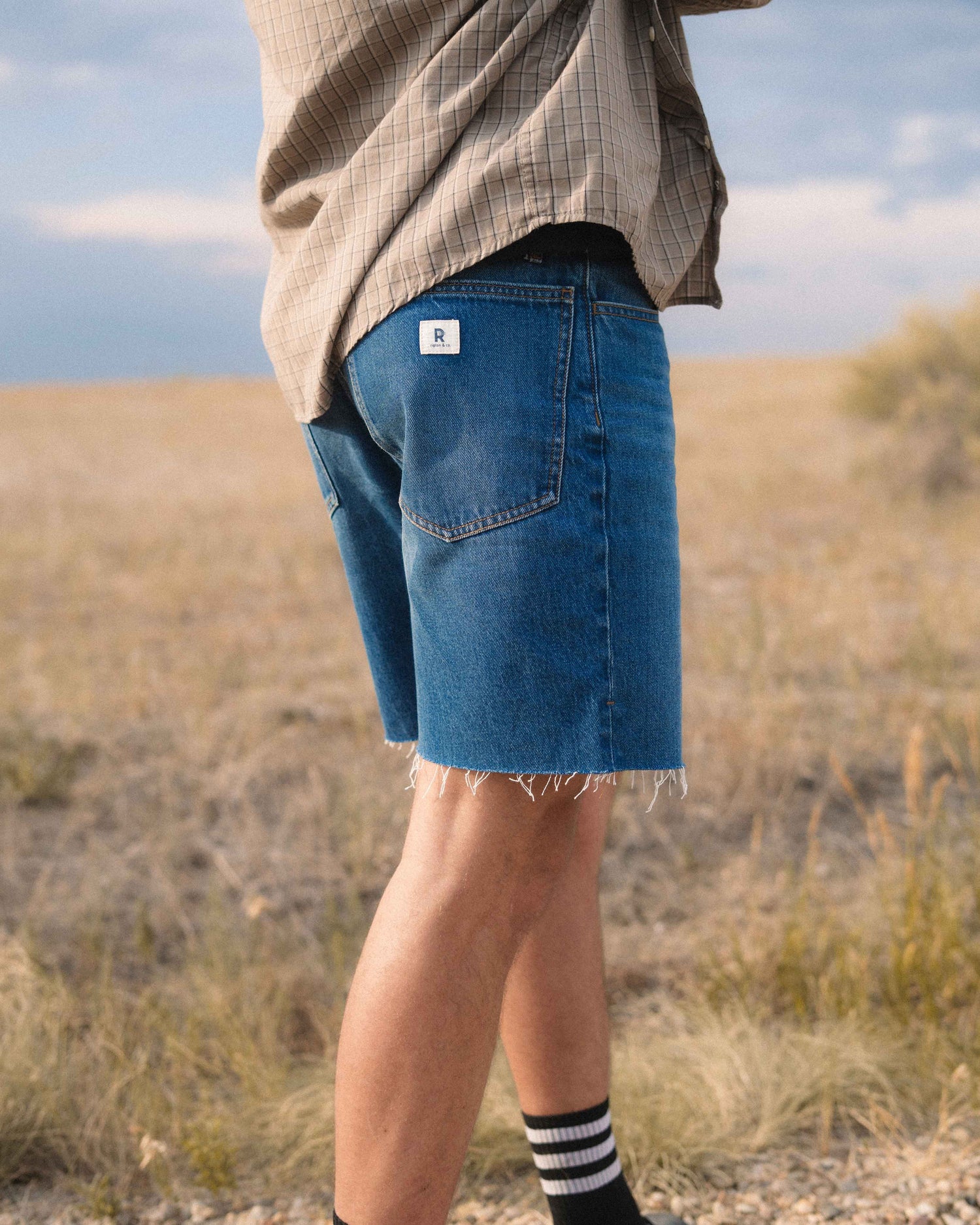 A person standing outdoors in a grassy field wearing blue denim shorts with a frayed hem, a beige overshirt, and black-and-white striped socks.