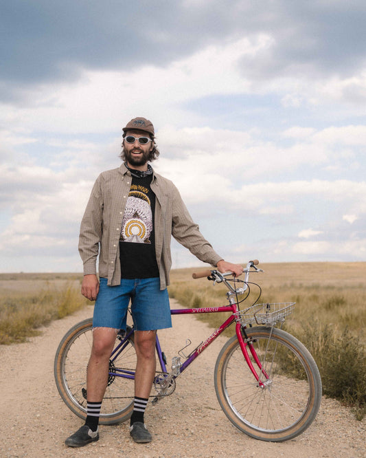 A man wearing a brown cap, sunglasses, a beige jacket, a black graphic t-shirt, and blue shorts stands on a dirt path next to a purple and red Schwinn bicycle under a cloudy sky.