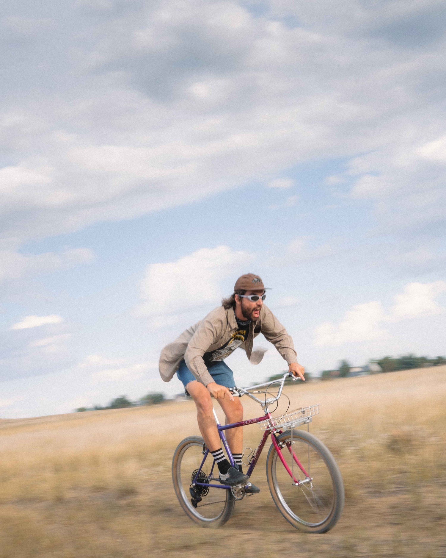 A man riding a bicycle across an open grassy field under a cloudy blue sky. The rider wears a tan jacket, denim shorts, sunglasses, and a brown beanie, and the bicycle has a purple frame with a silver basket.