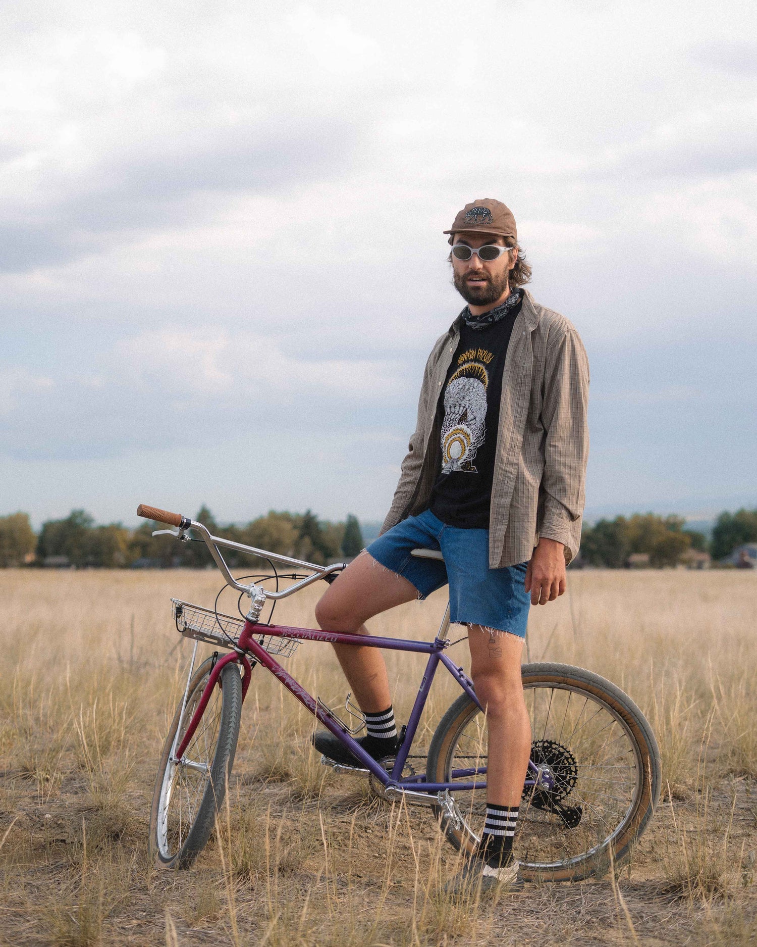 A man wearing a brown cap, sunglasses, a beige jacket, a black graphic t-shirt, and blue denim shorts stands next to a purple vintage bicycle in an open grassy field under a cloudy sky.