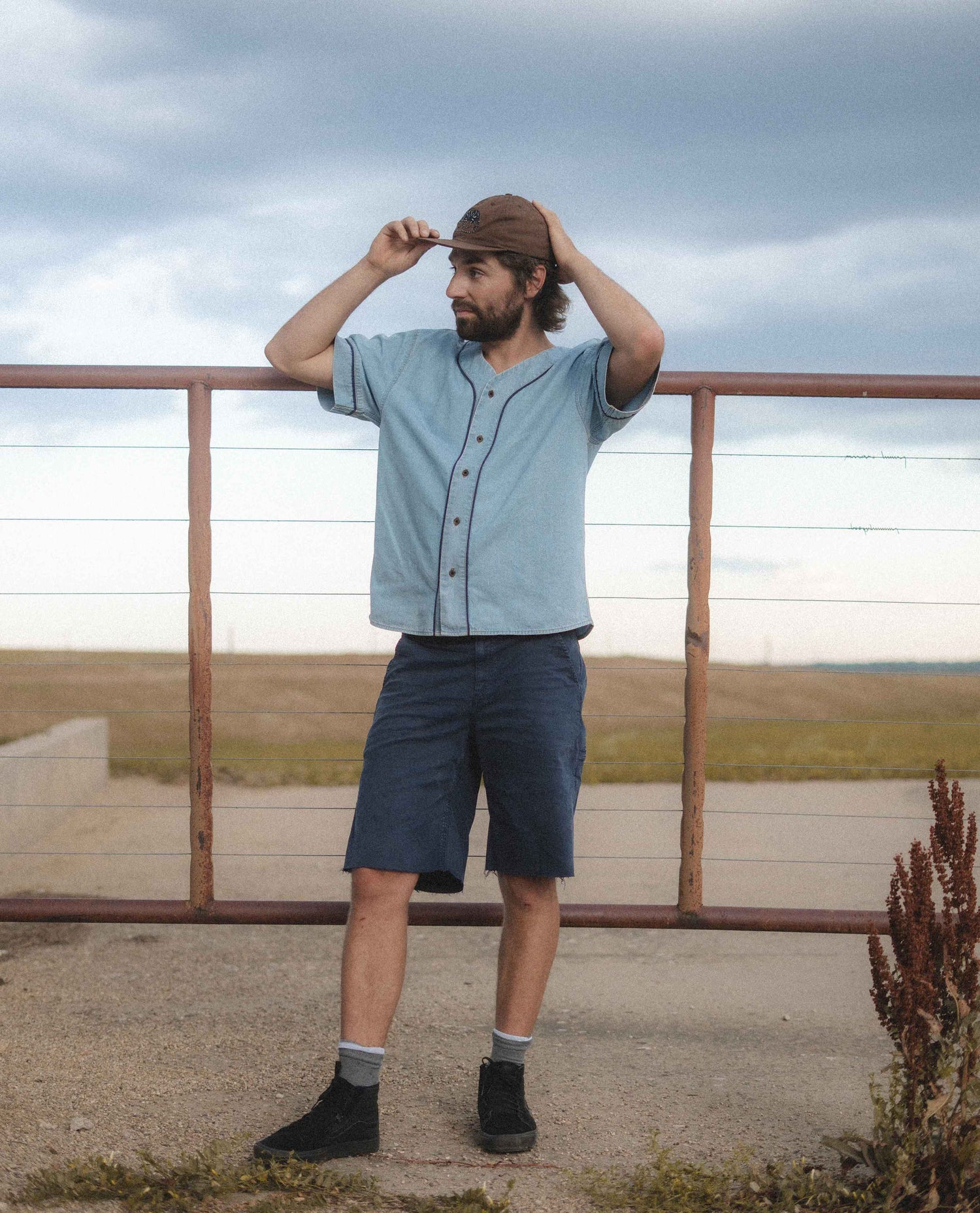 A man wearing a light blue short-sleeve button-up shirt, dark blue shorts, and a brown cap stands outdoors in front of a metal railing, looking upward under a cloudy sky.