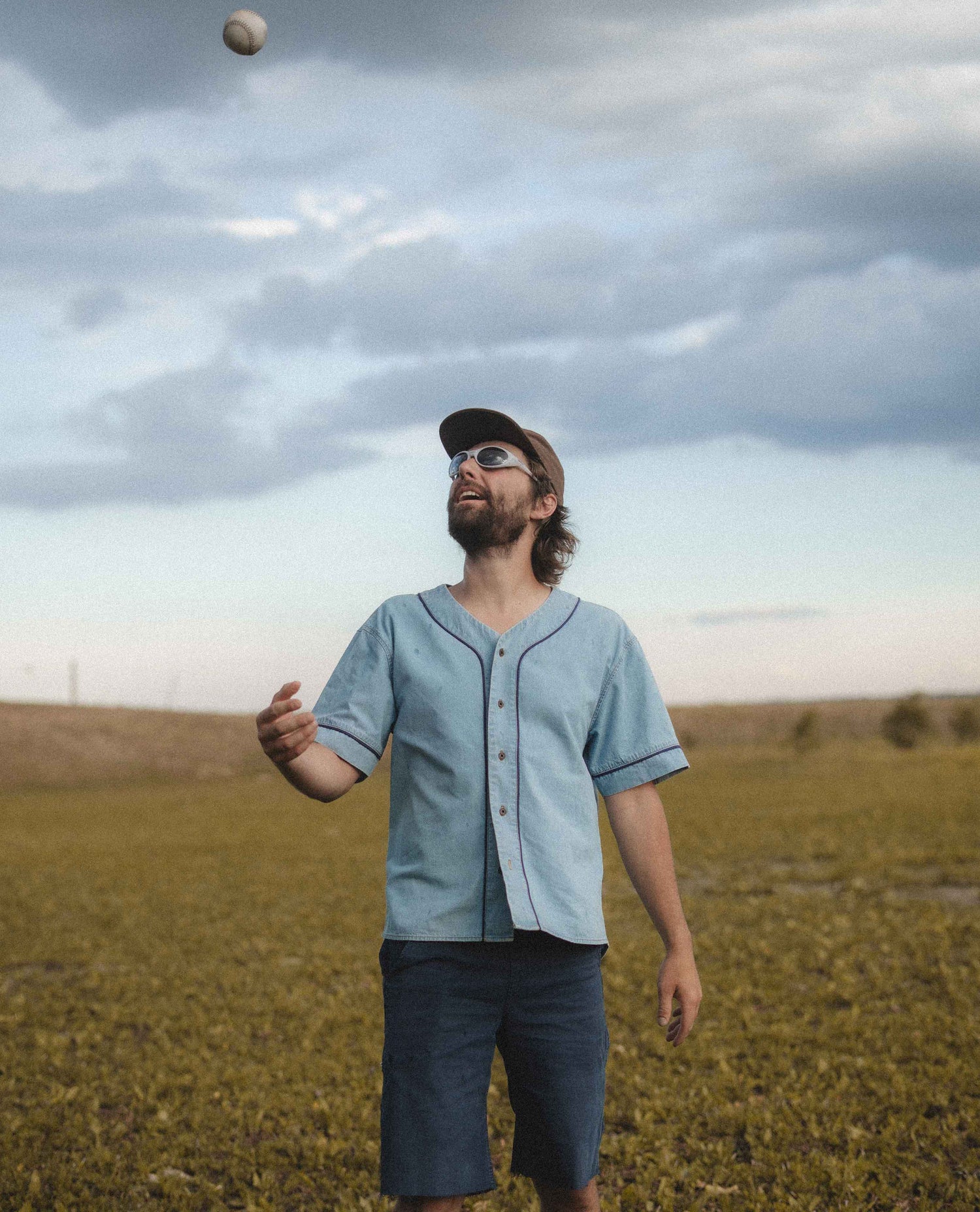 A man wearing a light blue short-sleeve button-up baseball shirt and dark shorts stands in an open field under a cloudy sky, looking up at a ball he is juggling.