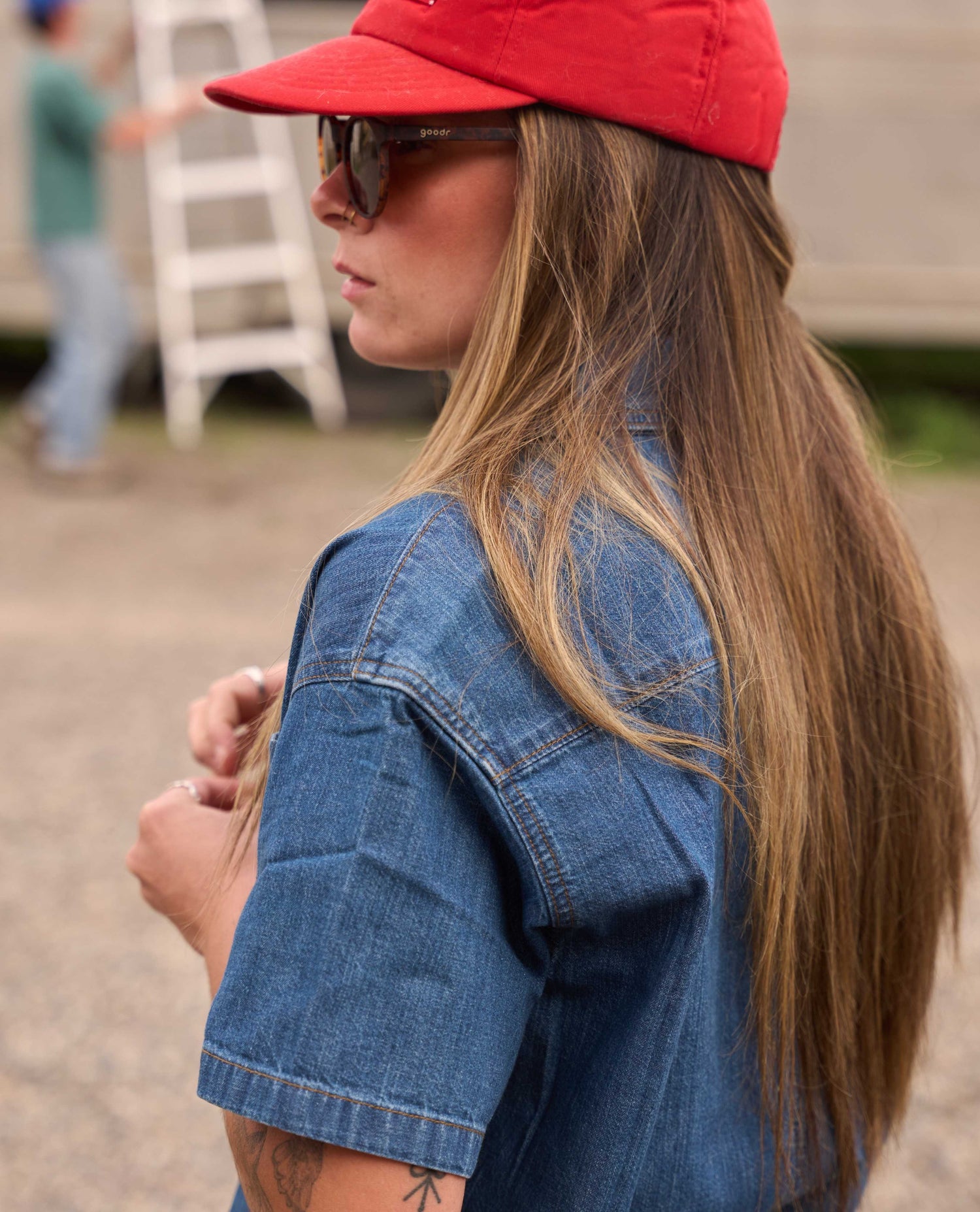A woman in profile wearing a red baseball cap, dark sunglasses, and a blue denim shirt, photographed outdoors with a blurred background.