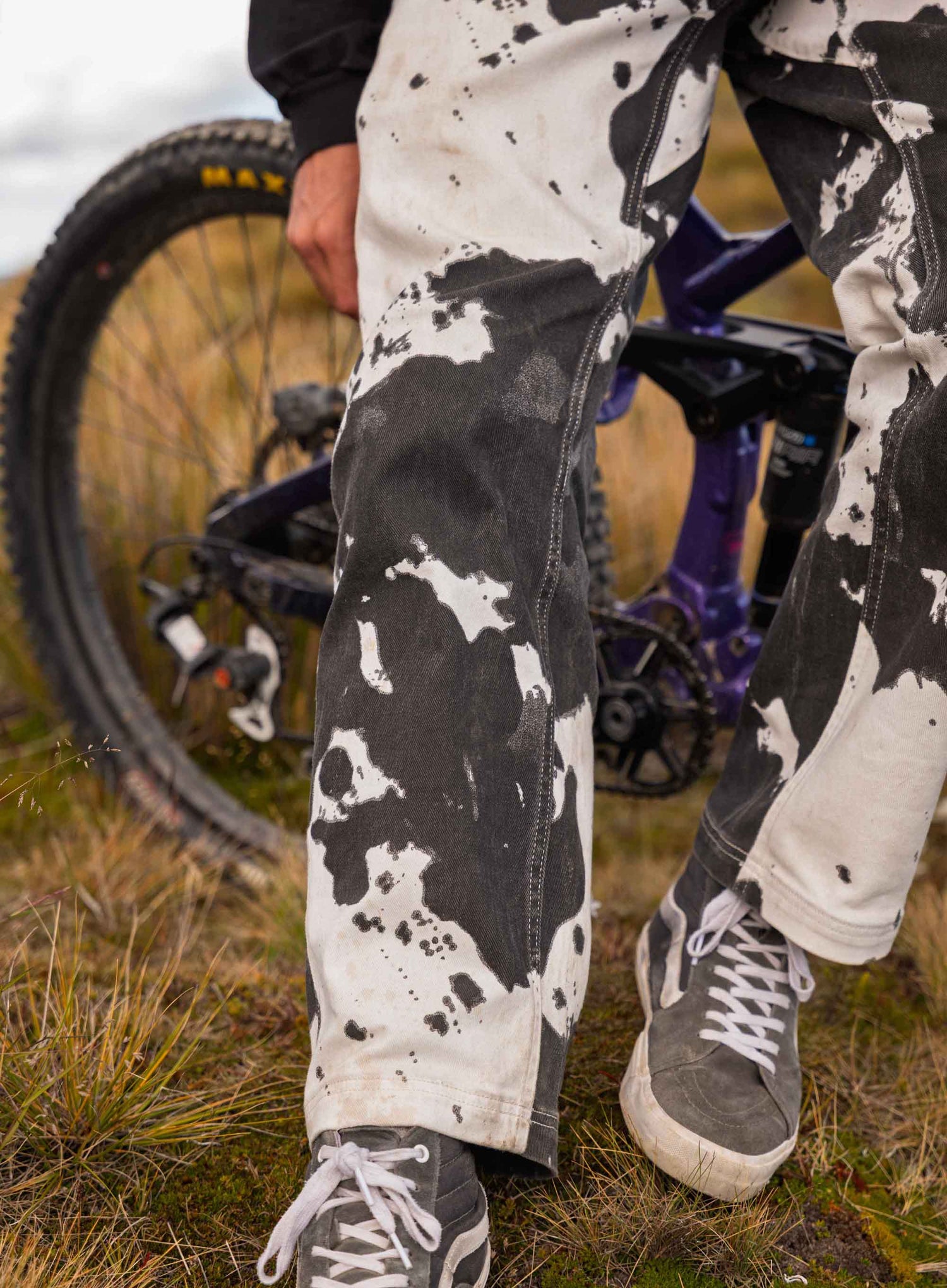 Person wearing white pants with black splatter pattern and gray sneakers standing next to a bicycle in an outdoor setting.