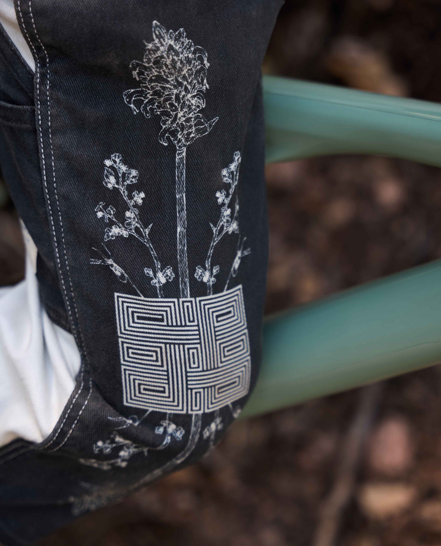 Close-up of a dark denim garment with white embroidered floral and geometric patterns, shown outdoors against a blurred natural background and a green metal structure.