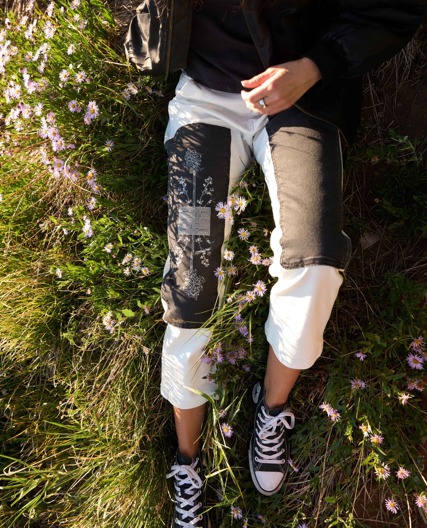 A person outdoors in a grassy field with small pink flowers, wearing white cropped pants with black patchwork knee panels, a black top, and black-and-white sneakers.