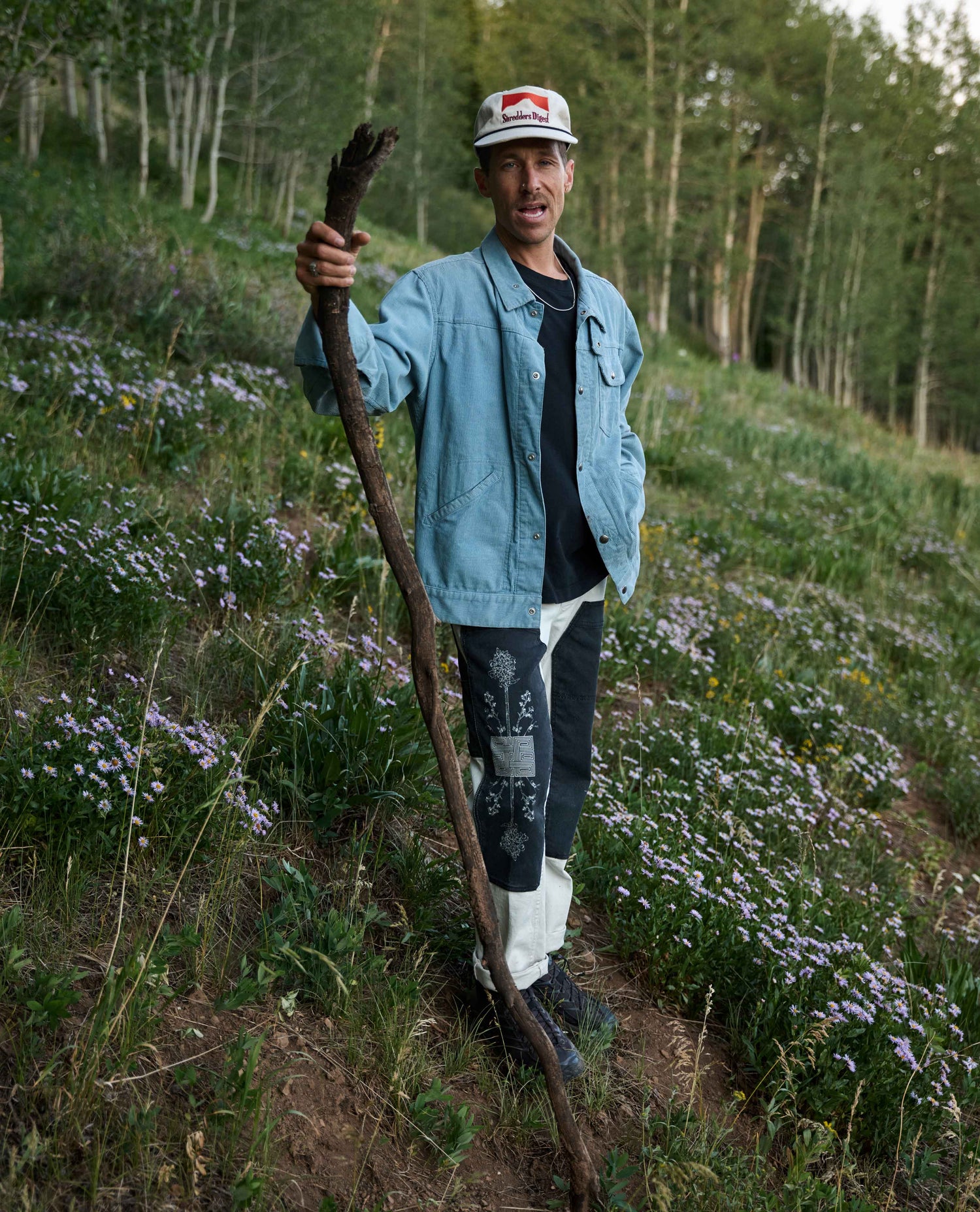A man standing outdoors on a grassy hill covered with small purple flowers, holding a long wooden staff. He is wearing a light blue denim jacket over a dark shirt, patterned pants, and a white cap with a red logo.