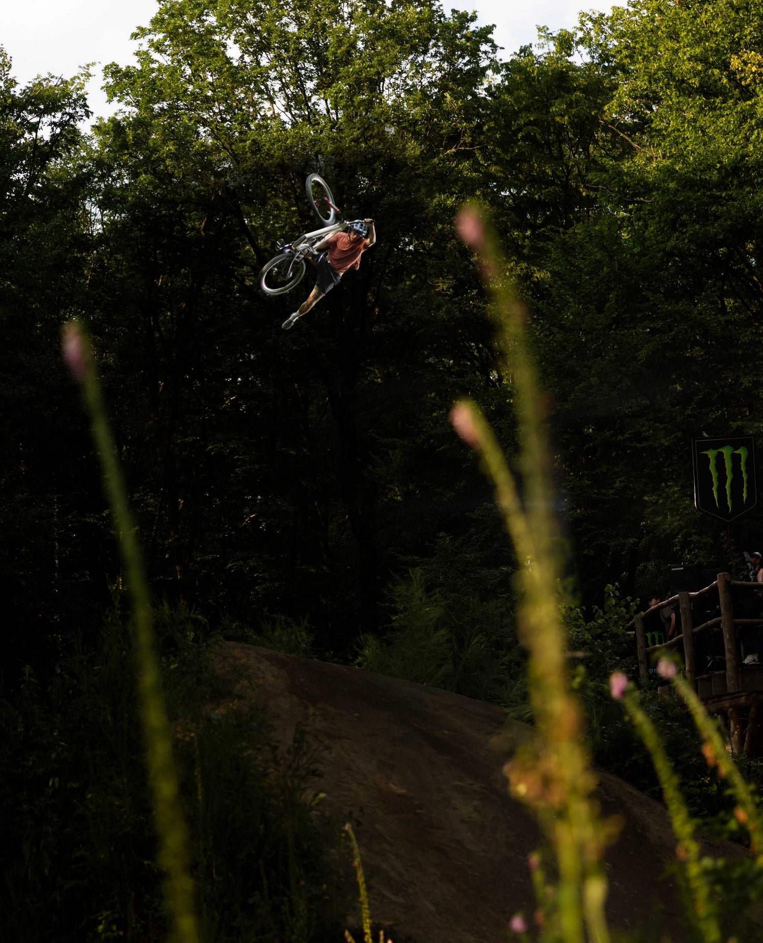 A mountain biker mid-air performing a jump trick in a dense forest, with blurred grass in the foreground and a dirt ramp below.