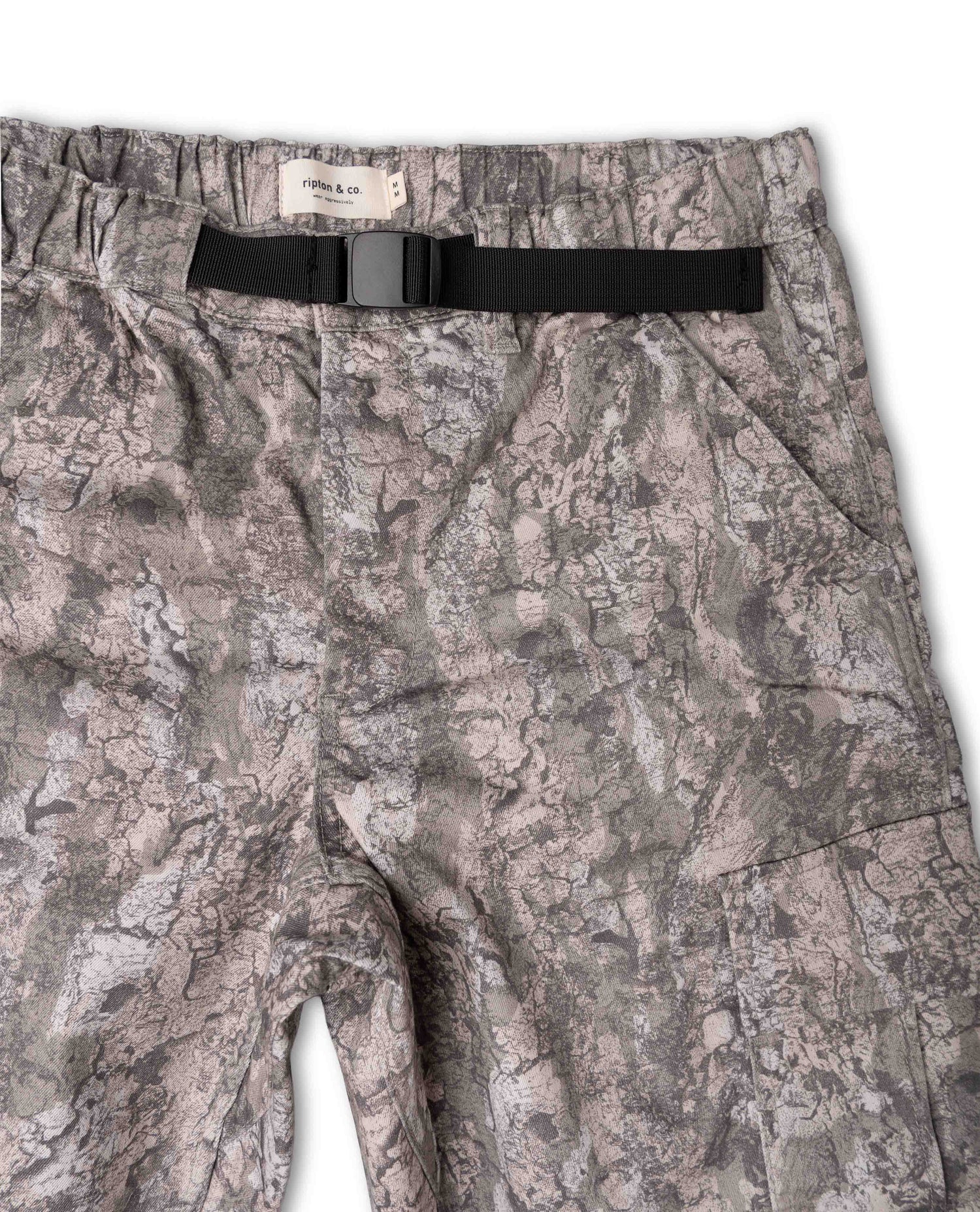 A close-up of a pair of camouflage shorts in a gray and beige woodland pattern, featuring a black belt with a silver buckle and a visible 'Ripton & CO' brand label at the waistband.