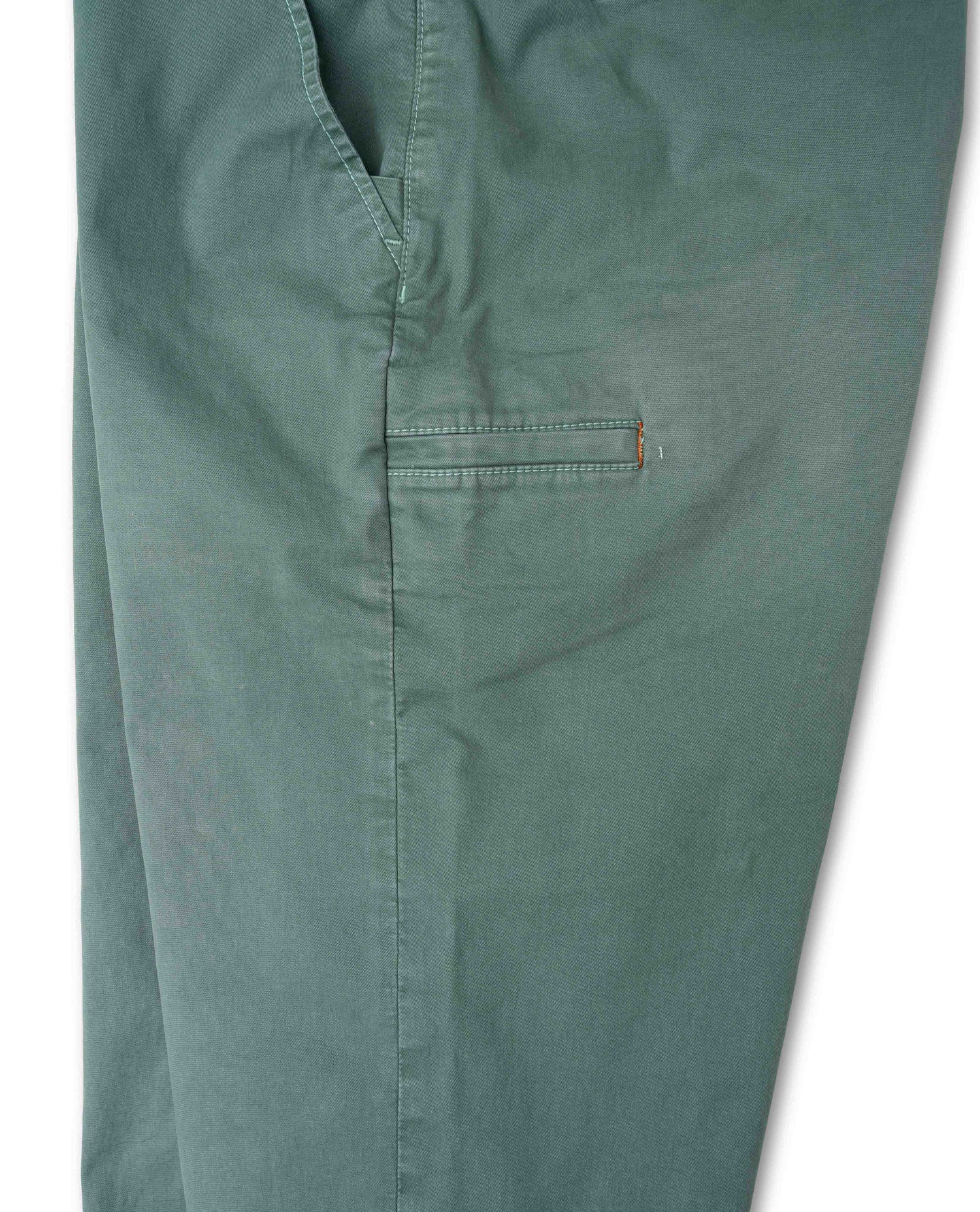 Close-up of a pair of muted sage green pants, showing the fabric texture, a vertical seam, and a small side pocket with a tiny orange tab accent against a white background.