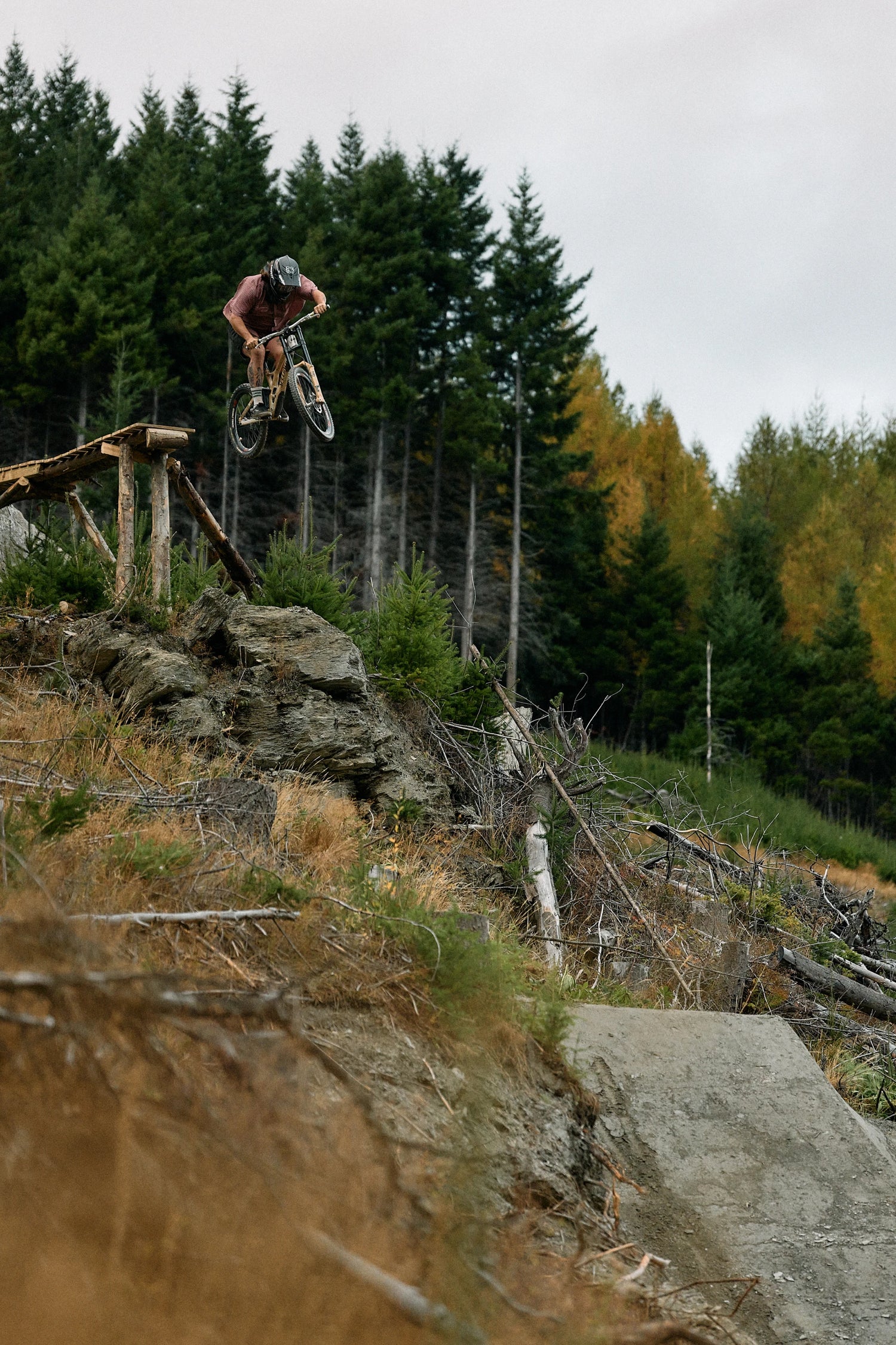 A mountain biker in a pink top and dark shorts jumps a bike over a wooden ramp on a dirt trail, with a forest background under an overcast sky.