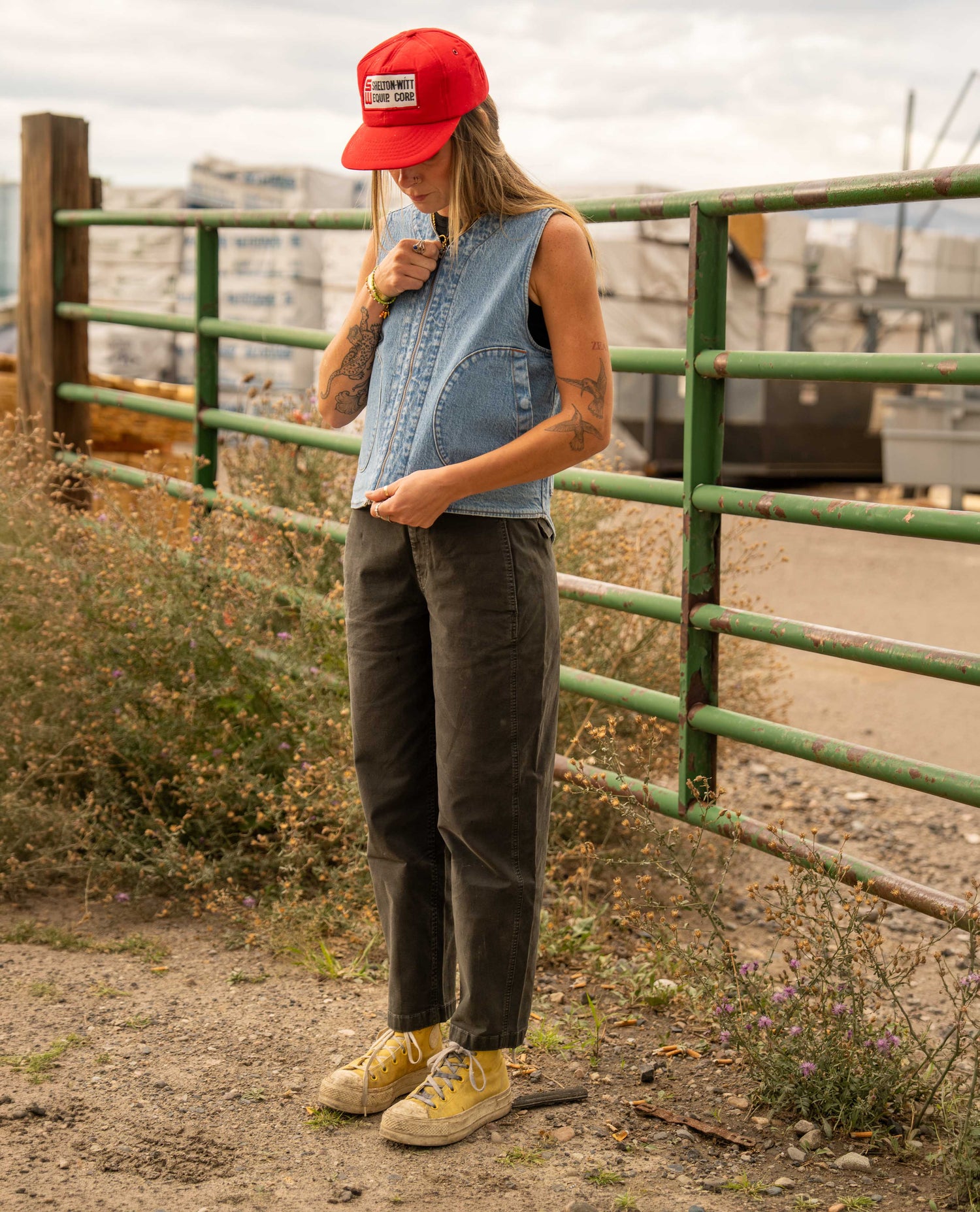 A person standing outdoors near a green metal fence, wearing a red baseball cap, a sleeveless light blue denim vest, dark olive pants, and yellow sneakers.