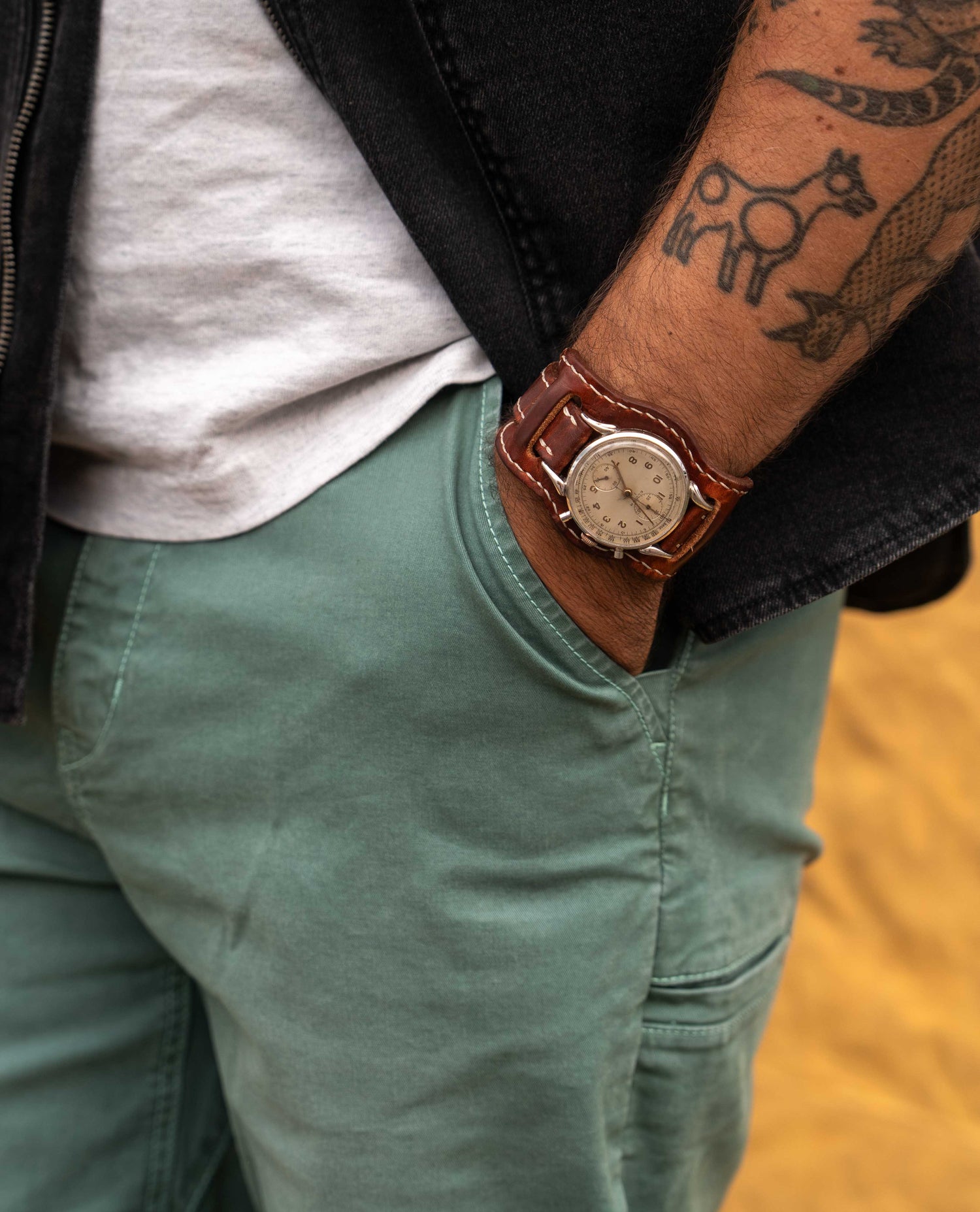 A close-up lifestyle shot of a man wearing a brown leather watch with a round beige dial, styled with a white t-shirt, black jacket, and sage green pants.