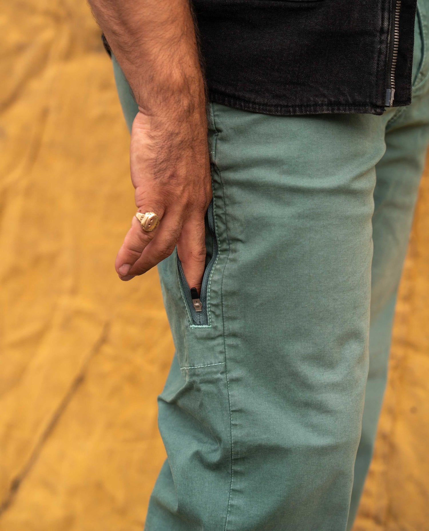 Close-up of a person wearing sage green work pants with a side zip pocket, shown with a hand reaching into the pocket. The person is also wearing a black denim jacket, and the background is a sandy, textured yellow-brown surface.