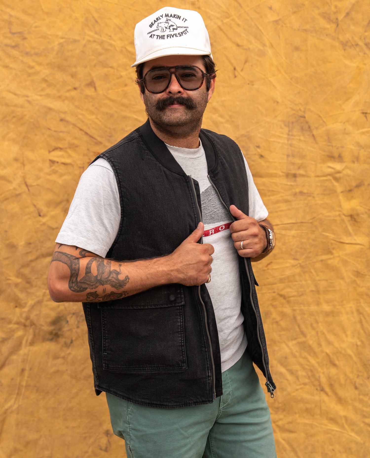 A man wearing a black denim vest over a white t-shirt, styled with a white cap, sunglasses, and light green pants, posing against a textured yellow backdrop.